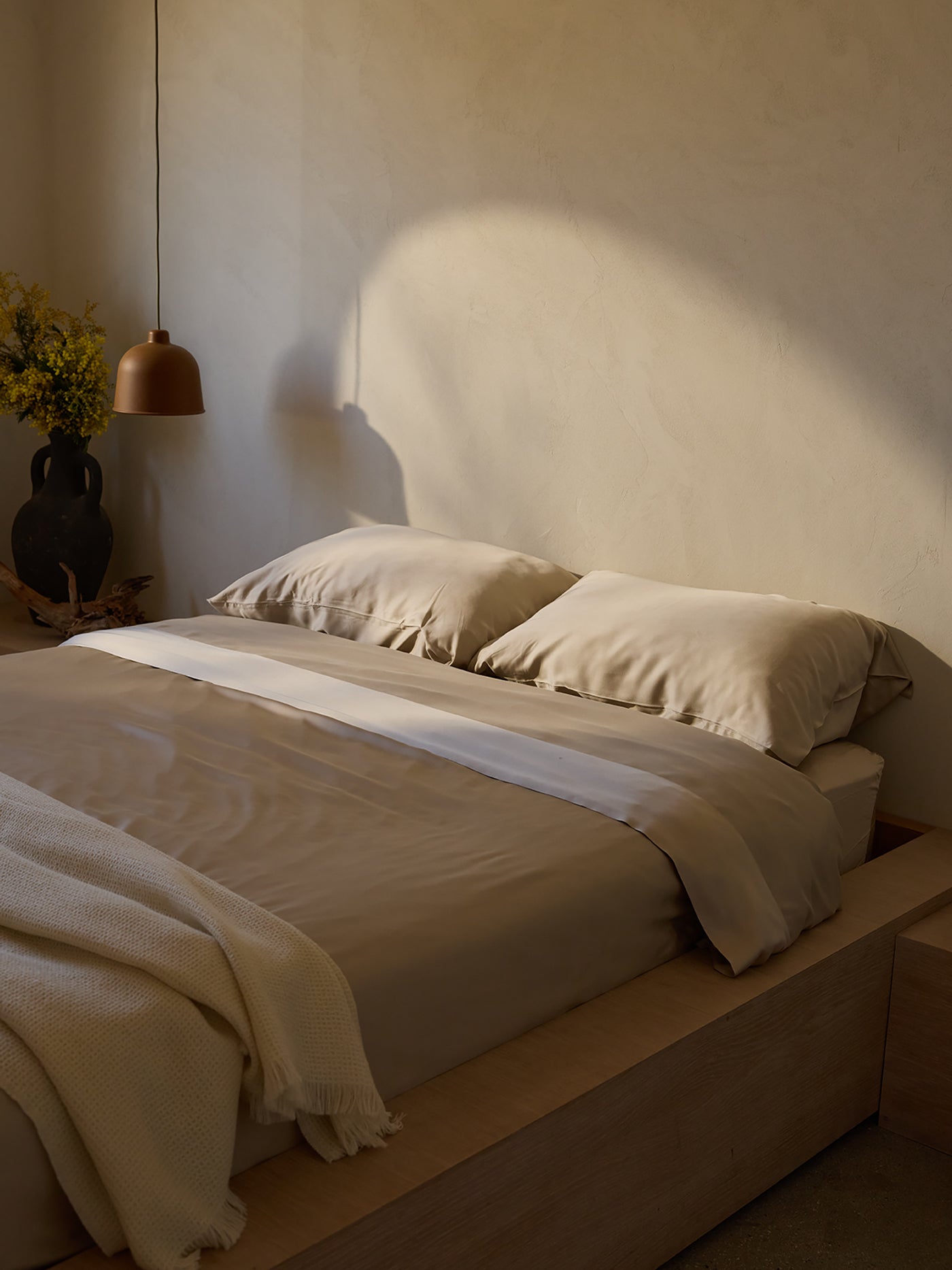 A minimalist bedroom featuring a wooden platform bed is adorned with Cozy Earth's Bamboo Sheet Set in light beige. Soft natural light creates shadows on the textured wall. A vase of yellow flowers decorates the bedside table, accompanied by a hanging bronze lamp. |Color:Driftwood