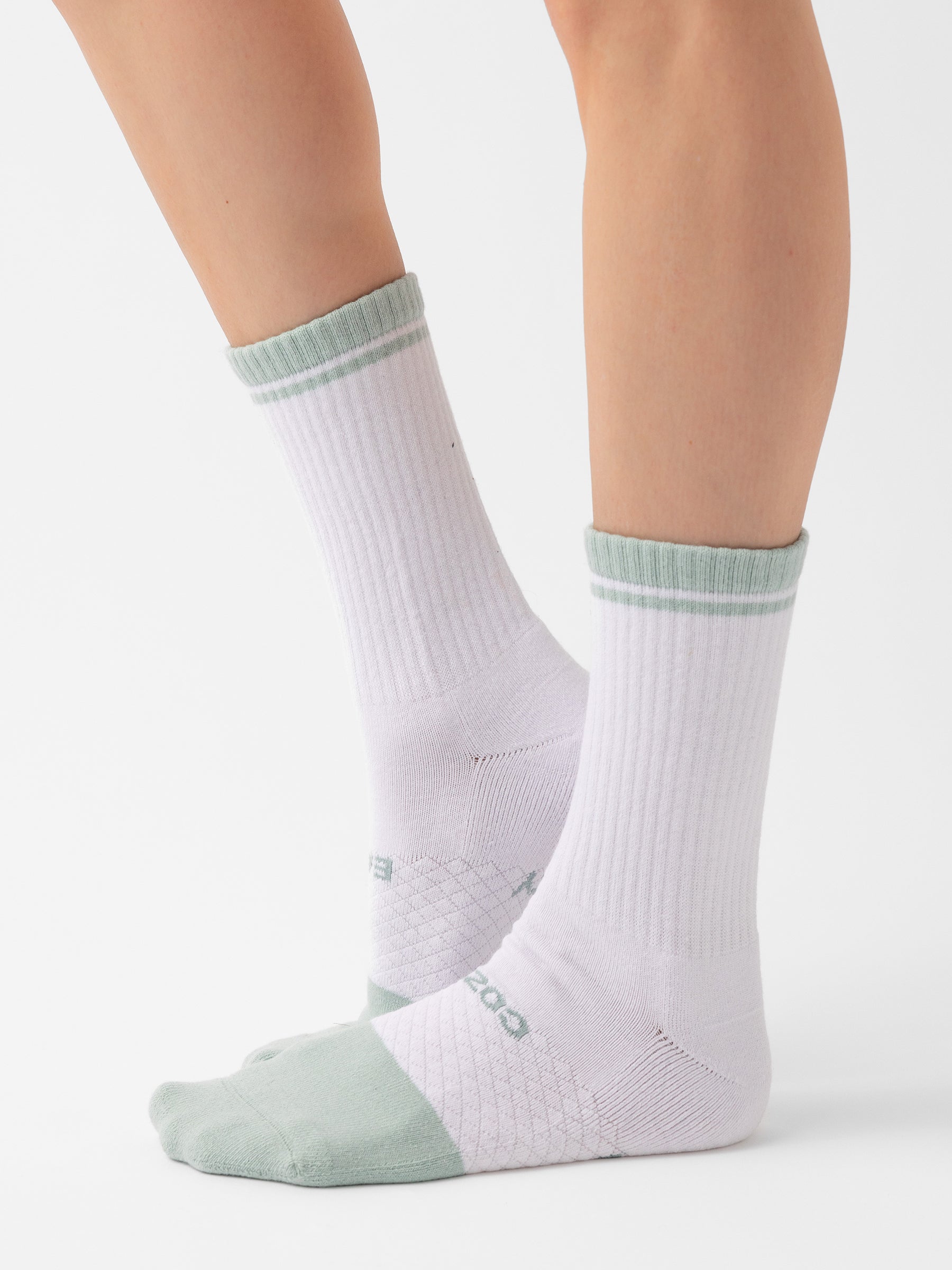 A close-up of a person wearing the "Essential Calf Sock 2-Pack" from Cozy Earth, featuring white crew socks with light green toes, heels, and a green stripe at the top. The mid-calf length socks have a ribbed texture. The person is standing against a plain white background. |Color:Arctic