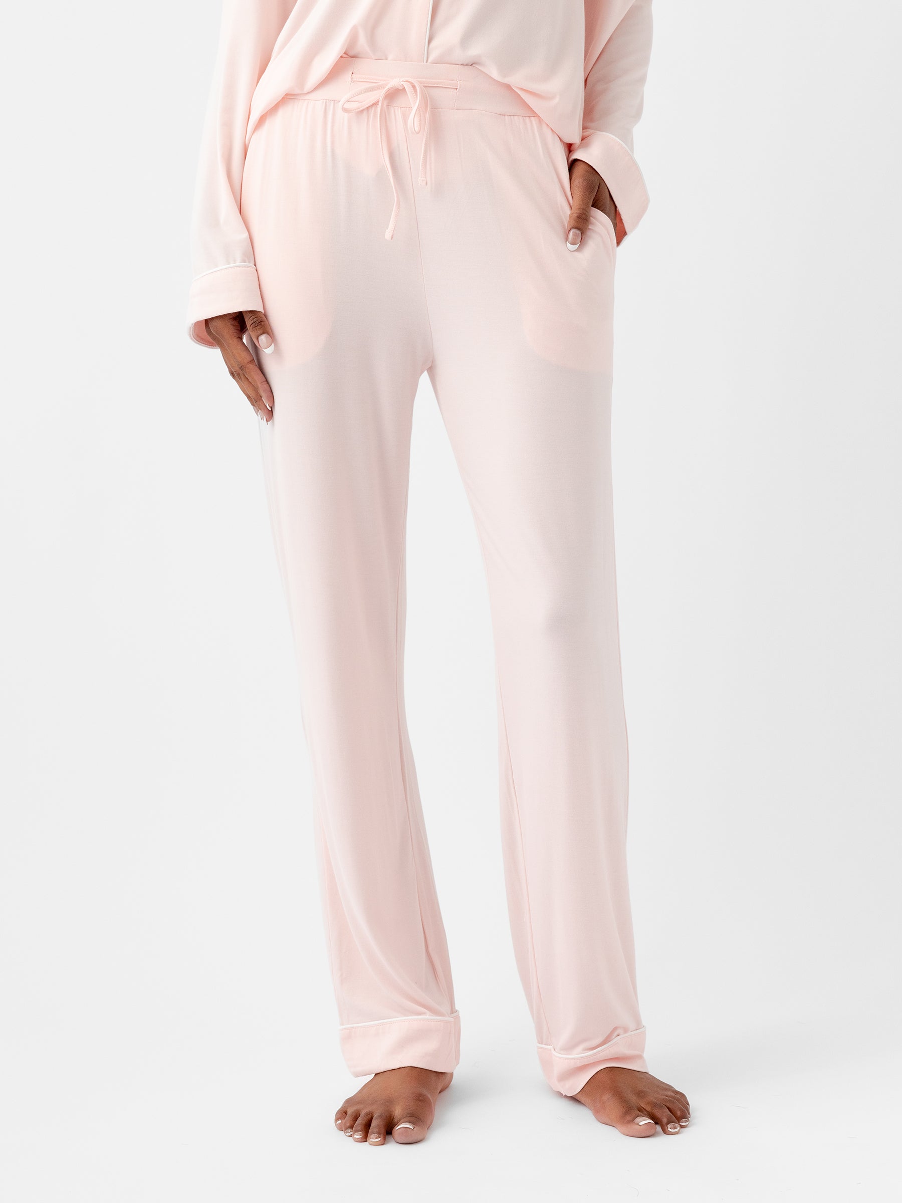 A woman wearing HIDE Women's Stretch-Knit Bamboo Pajama Pants in light pink stands with one hand in her pocket. The loose-fitting pants feature an elastic waistband and a drawstring tie. Her bare feet are visible against a plain white background. |Color:Blossom