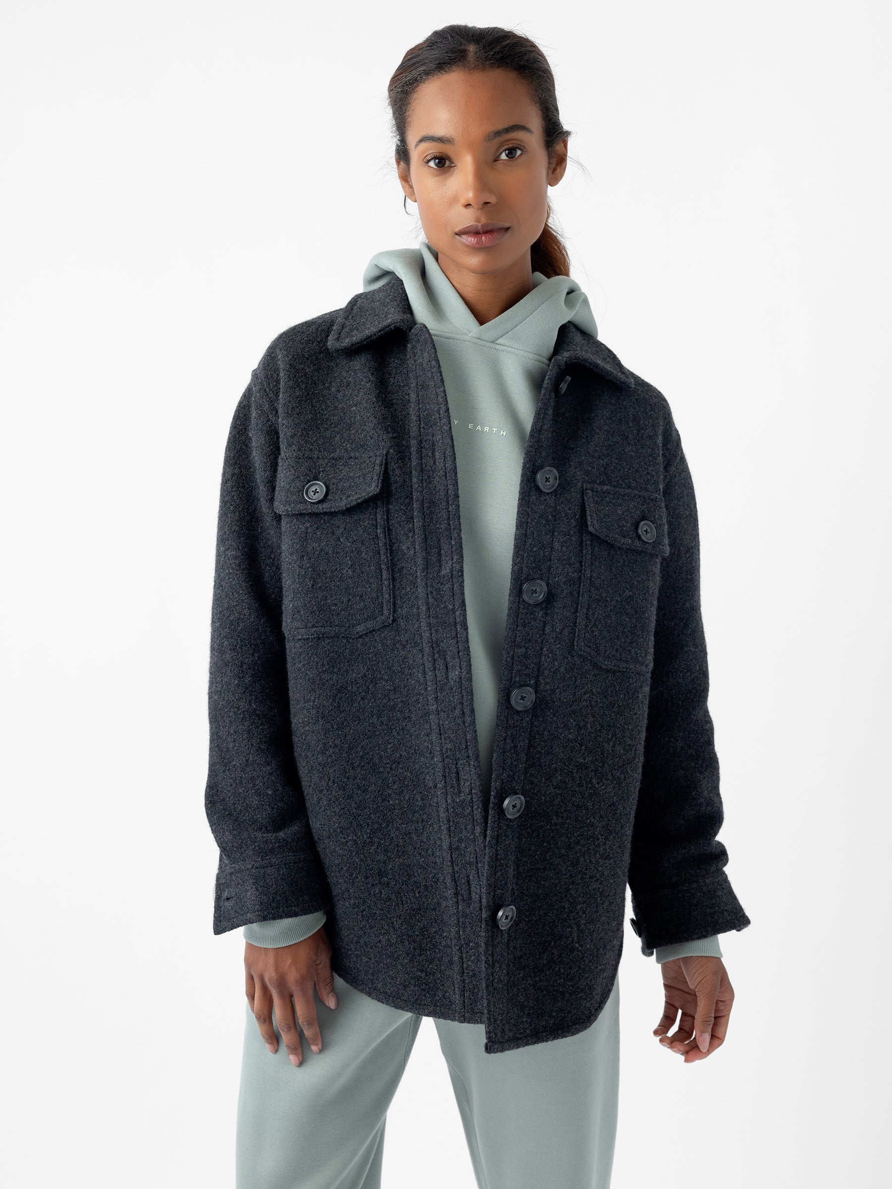 A person with long hair tied back is wearing the Women's Boucle Shacket by Cozy Earth, buttoned up over a light gray hoodie. They are standing against a plain white background, looking directly at the camera with a neutral expression. |Color:Coal