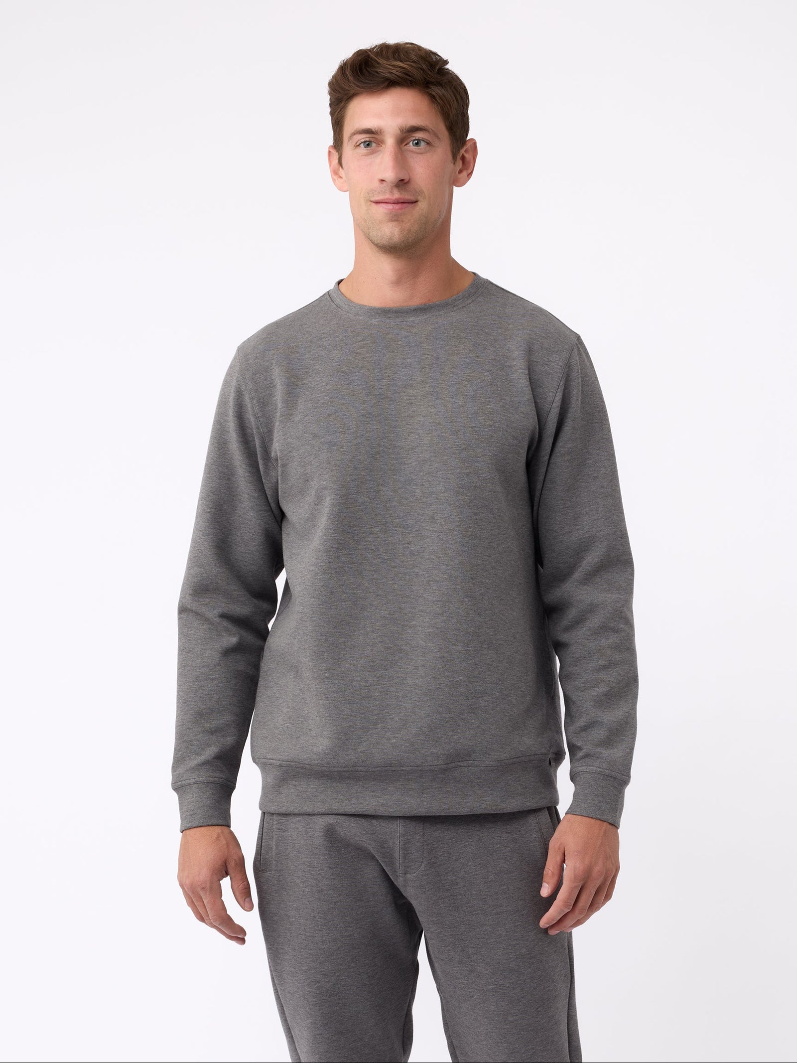 A man is wearing the Men's StretchTech Crewneck sweatshirt from Cozy Earth, paired with matching sweatpants. He stands against a plain white background with short brown hair, looking forward with a neutral expression. 