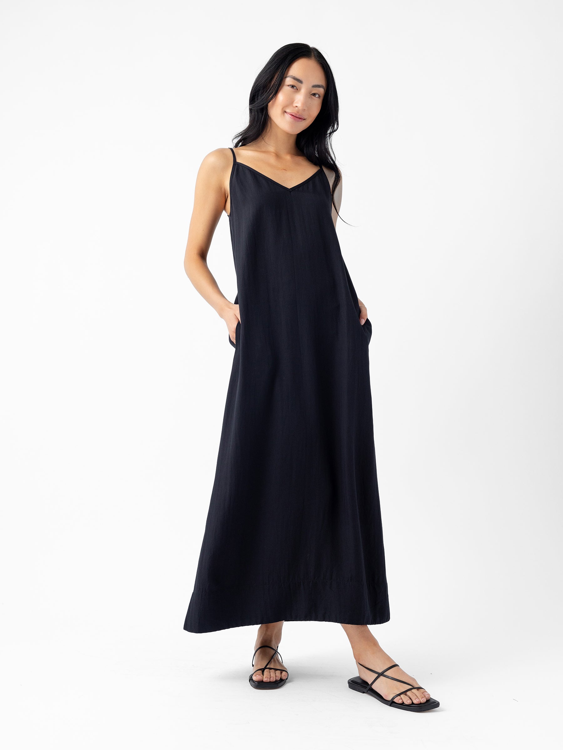 A person with long, dark hair wearing the Cozy Earth Women's Sunset Slip Dress stands against a plain white background. They wear black strappy sandals and have their hands in the dress's side pockets, smiling gently. |Color:Jet Black