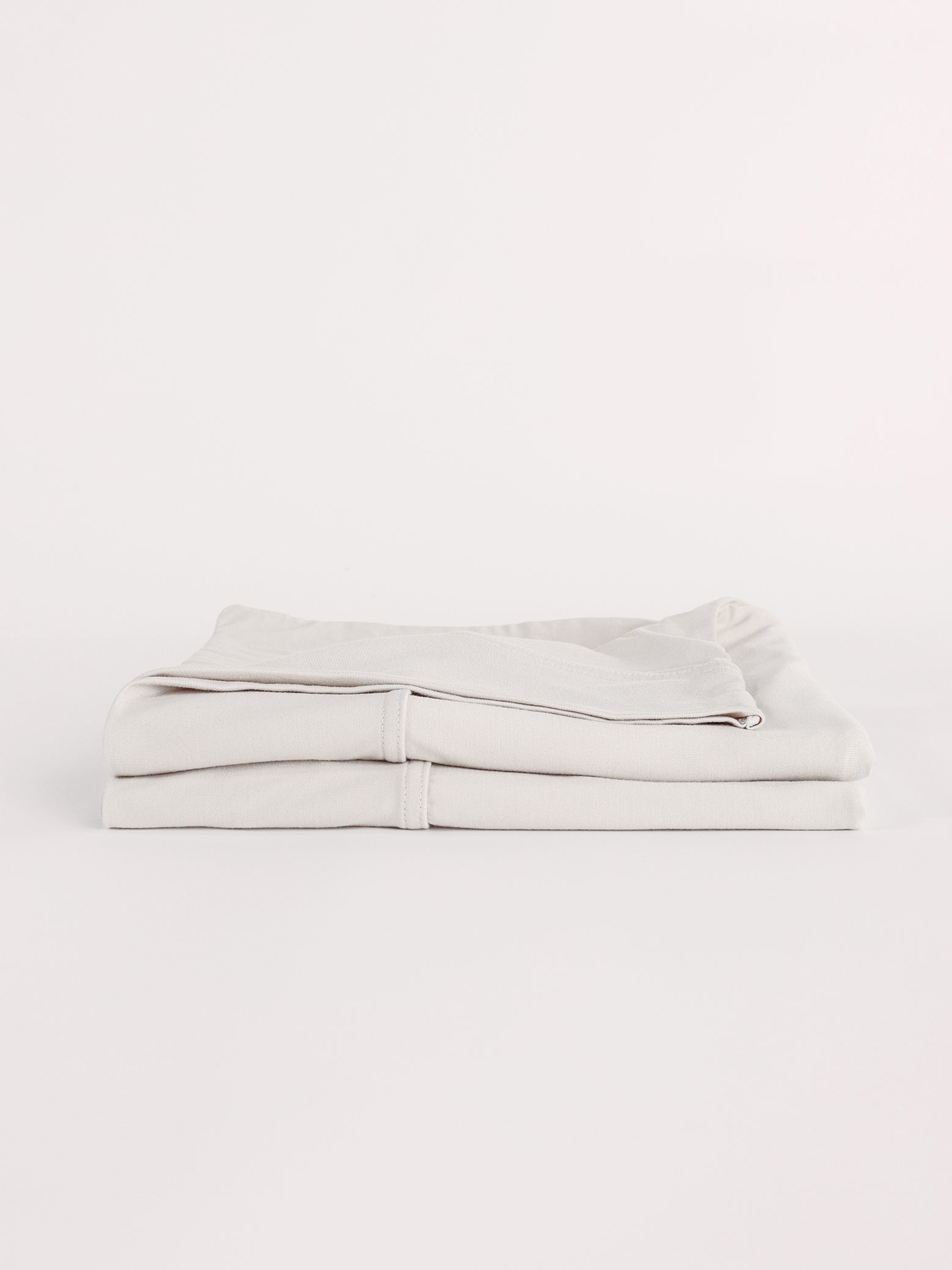 A neatly folded Bamboo Jersey Flat Sheet by Cozy Earth is displayed against a white background. The light gray sheets are tightly stacked, showcasing their smooth texture and clean appearance. |Color:Light Grey