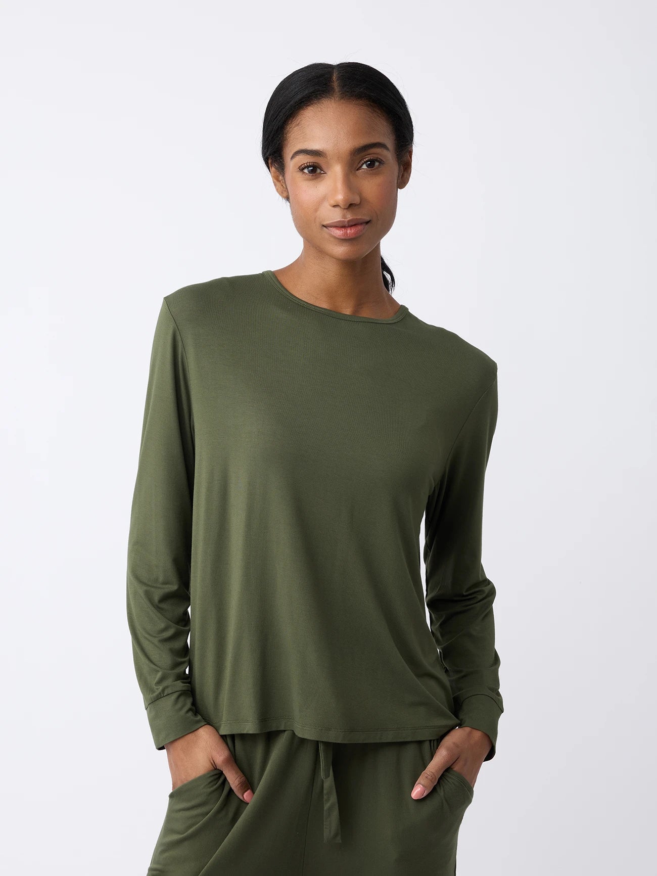 A woman stands against a white background wearing the Women's Stretch Knit Bamboo Long Sleeve Lounge Tee by Cozy Earth, paired with matching pants. They have long, dark hair pulled back and smile softly with hands in their pockets. |Color:Olive