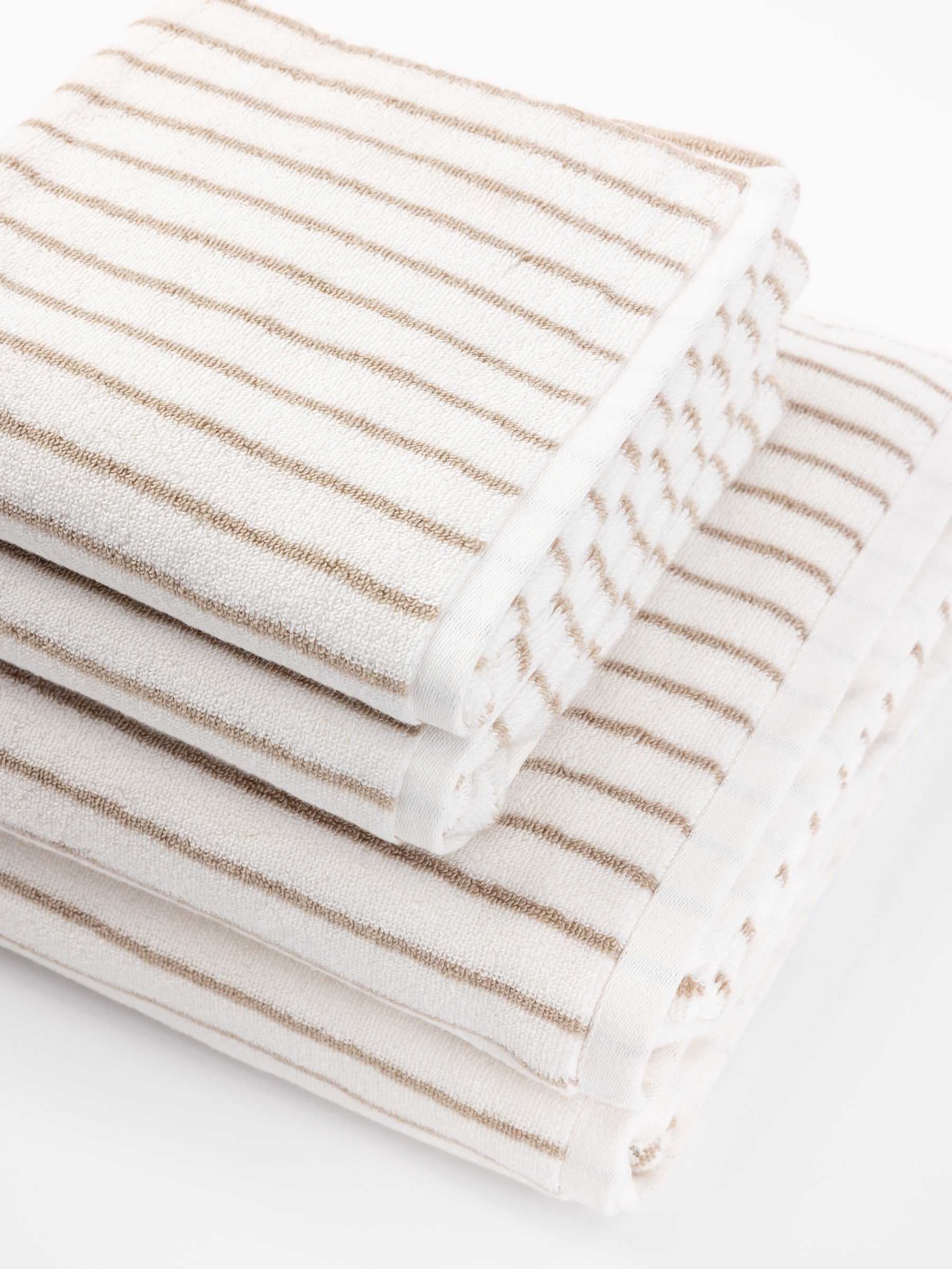 A stack of three Luxe Bath Towels by Cozy Earth in white with light brown stripes are neatly arranged, with each towel slightly offset from the one beneath it. The simple, elegant design gives a clean and inviting appearance. |Color:Sand Stripe