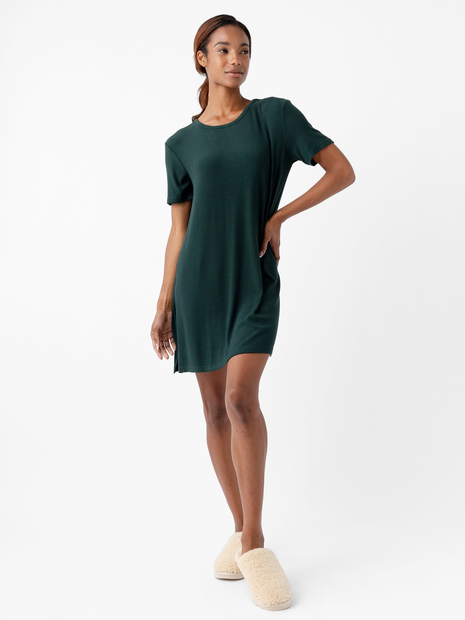A person wearing a Cozy Earth Women’s Bamboo Rib-Knit Boyfriend Sleep Dress in dark green and light-colored, fuzzy slippers stands against a plain white background. The individual has one hand resting on their hip and is looking confidently into the distance. 