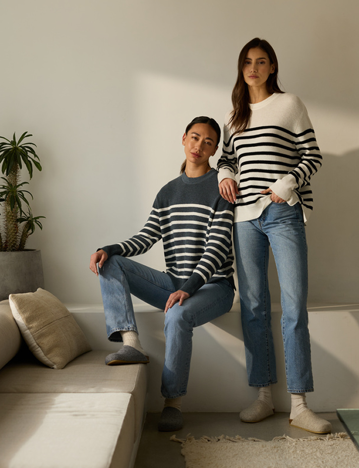Two people in a softly lit room wearing striped sweaters and jeans. One is seated on a couch, and the other stands beside them. There's a small potted plant on a table, and the ambiance is calm and casual.