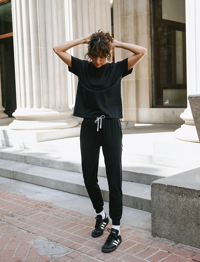 A person with curly hair stands on a sidewalk with their hands on their head, wearing a black t-shirt, black joggers, and black sneakers. They are in front of a building with large columns and steps.