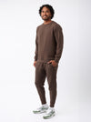 A man with curly hair smiles against a white background, wearing the Cozy Earth Men's StretchTech Crewneck & Jogger Set in brown, paired with gray-and-white sneakers. |Color:Cacao