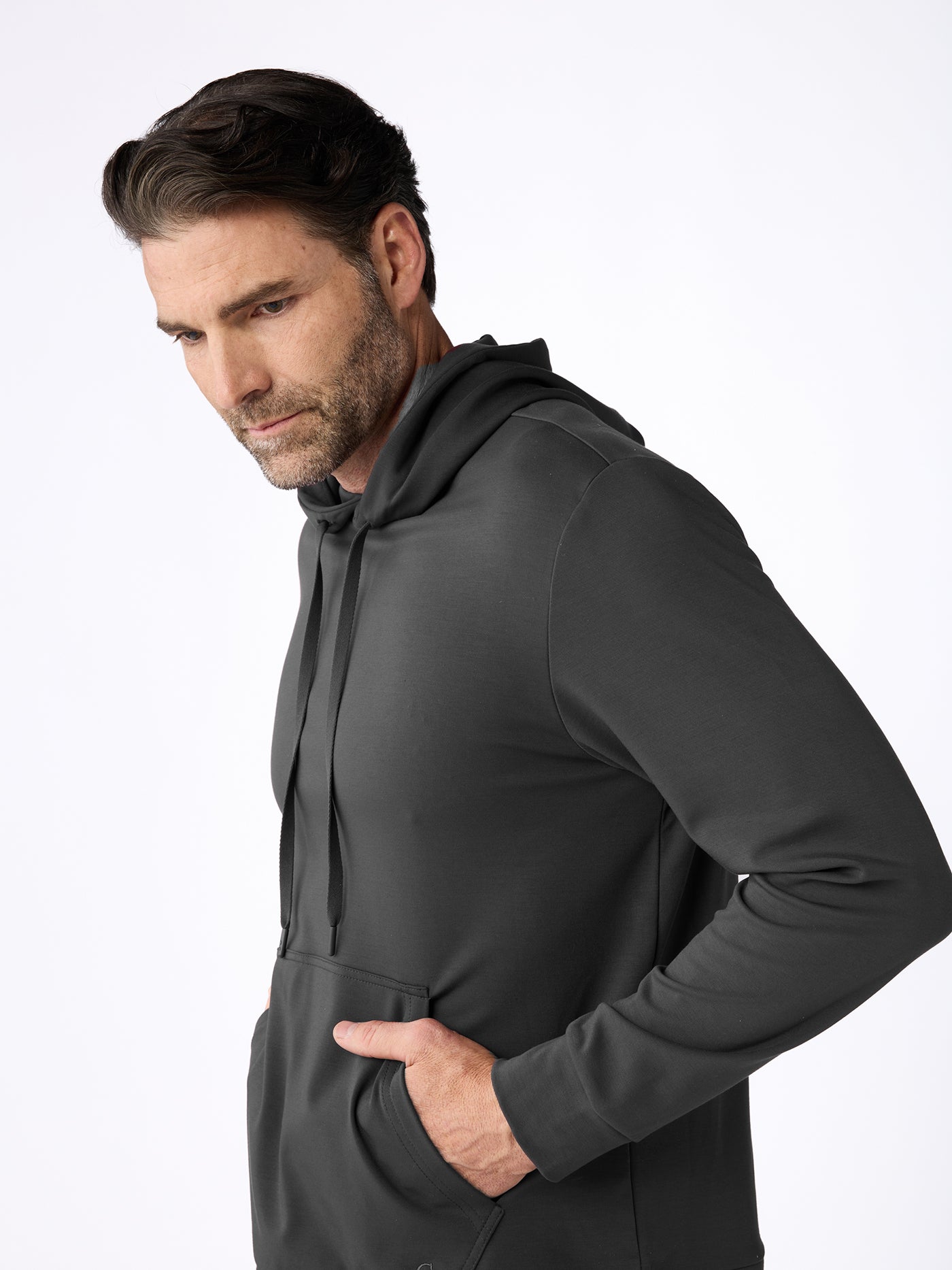 A man with dark hair and a beard wears the Cozy Earth Men's Brushed Bamboo Hoodie & Jogger Set in dark gray, standing with one hand in his pocket and looking slightly downward against a plain white background. 