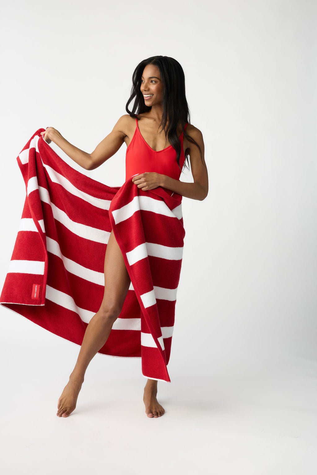 A woman in a red swimsuit holds the Cozy Earth Classic Stripe Resort Towel, dressed in striking red and white stripes, partially wrapped around her. She stands barefoot against a plain white backdrop, smiling and looking to her right. |Color:Crimson