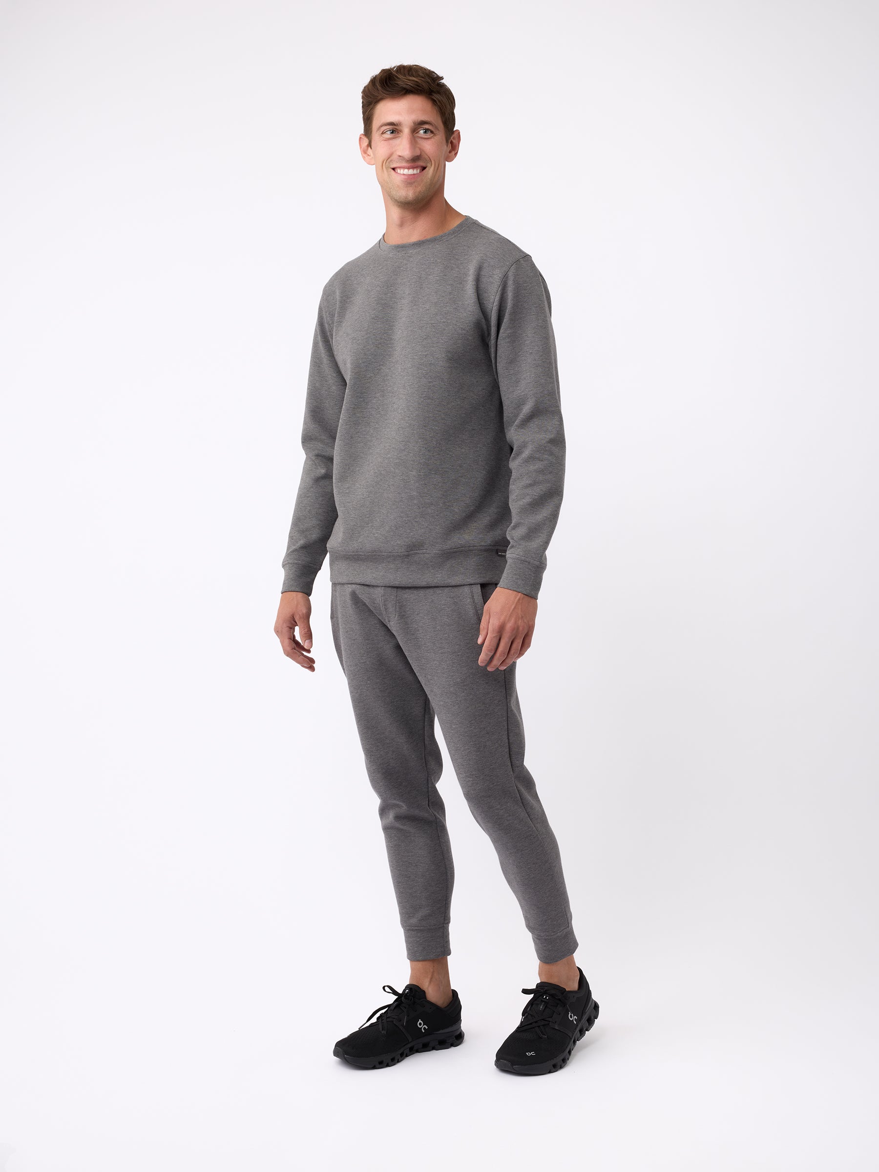 A man is smiling and wearing a Cozy Earth matching gray Men's StretchTech Crewneck sweatsuit with black sneakers. He is standing against a plain white background. |Color:Coal Heather