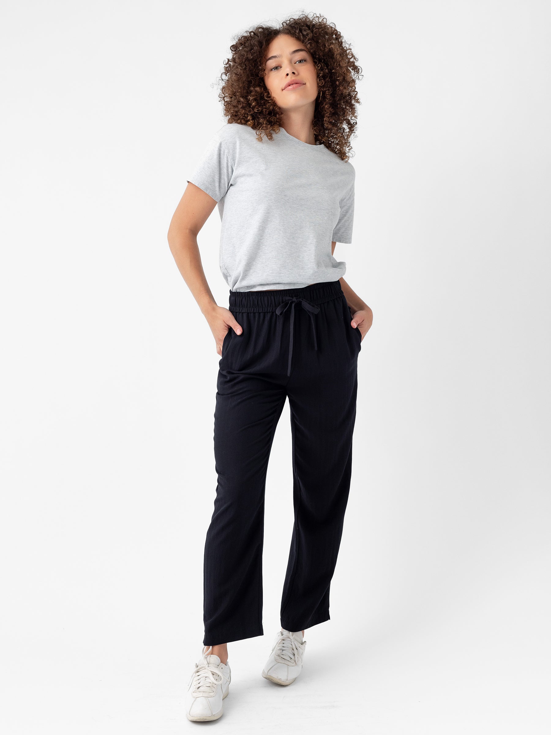 A person with curly hair is standing against a white background. They are wearing a light gray T-shirt, Cozy Earth's Women's Sunset Cropped Pants, and white sneakers. Their hands are in their pockets, and they have a relaxed expression on their face. |Color:Jet Black