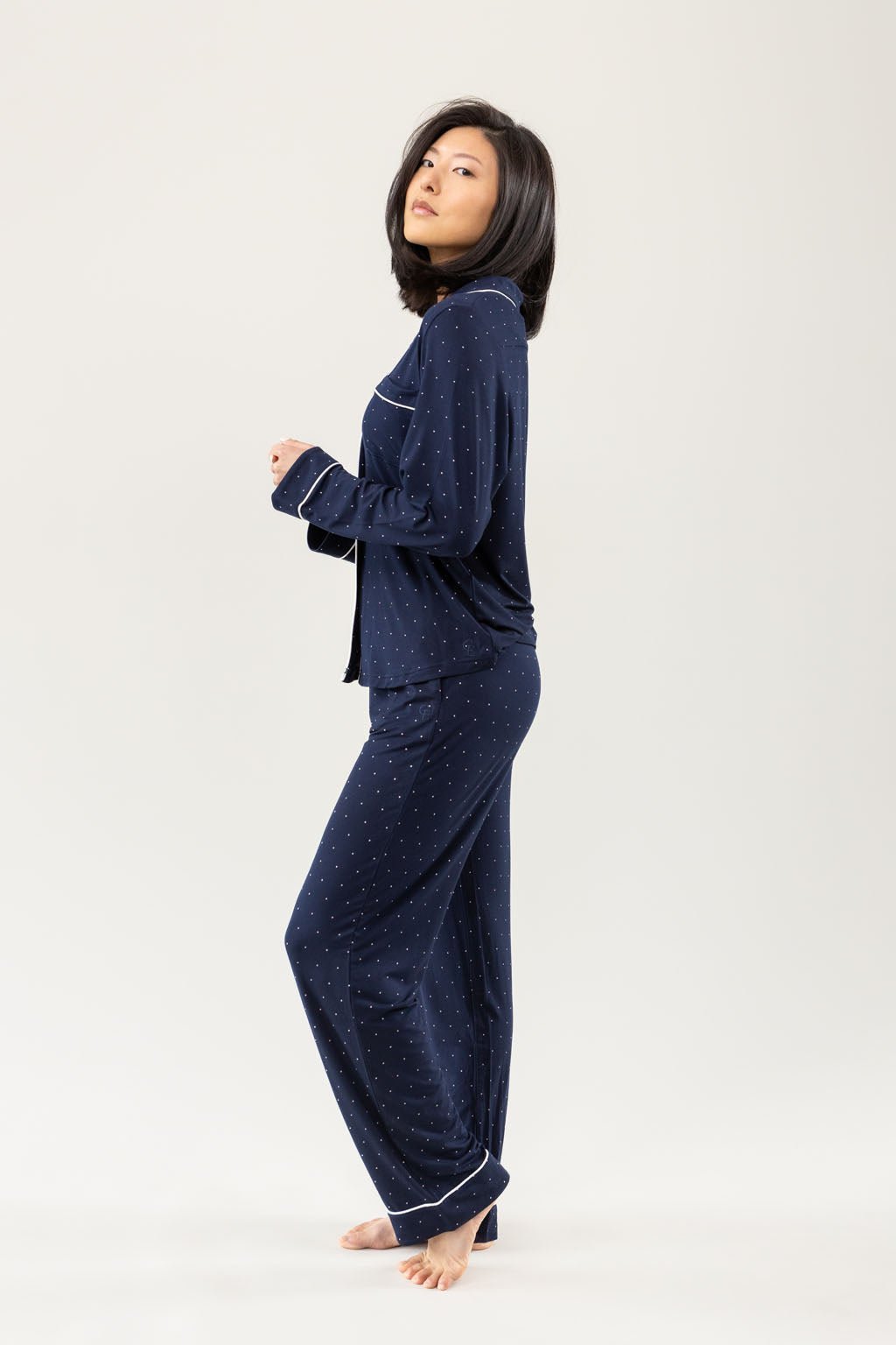 A person with shoulder-length hair stands barefoot, wearing Cozy Earth's Women's Bamboo Stretch-Knit Long Sleeve Pajama Set in dark blue with white dotted patterns, posed in profile against a plain background. 