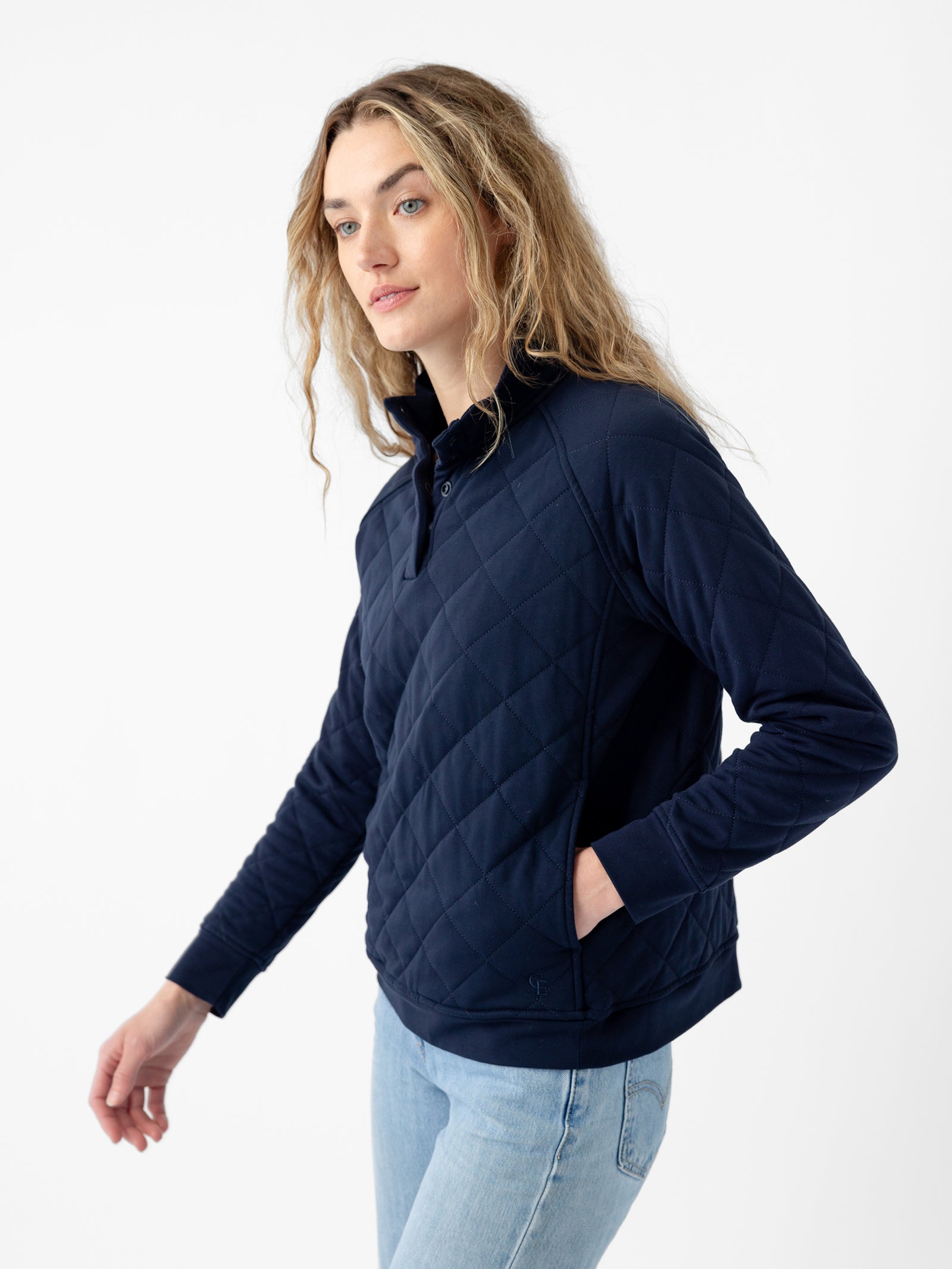 A person with long, wavy hair wearing the Cozy Earth Women's Ultra-Soft Bamboo Quilted Snap Pullover in dark blue and light blue jeans stands against a plain white background. They are looking slightly off to the side with one hand in a pullover pocket. 
