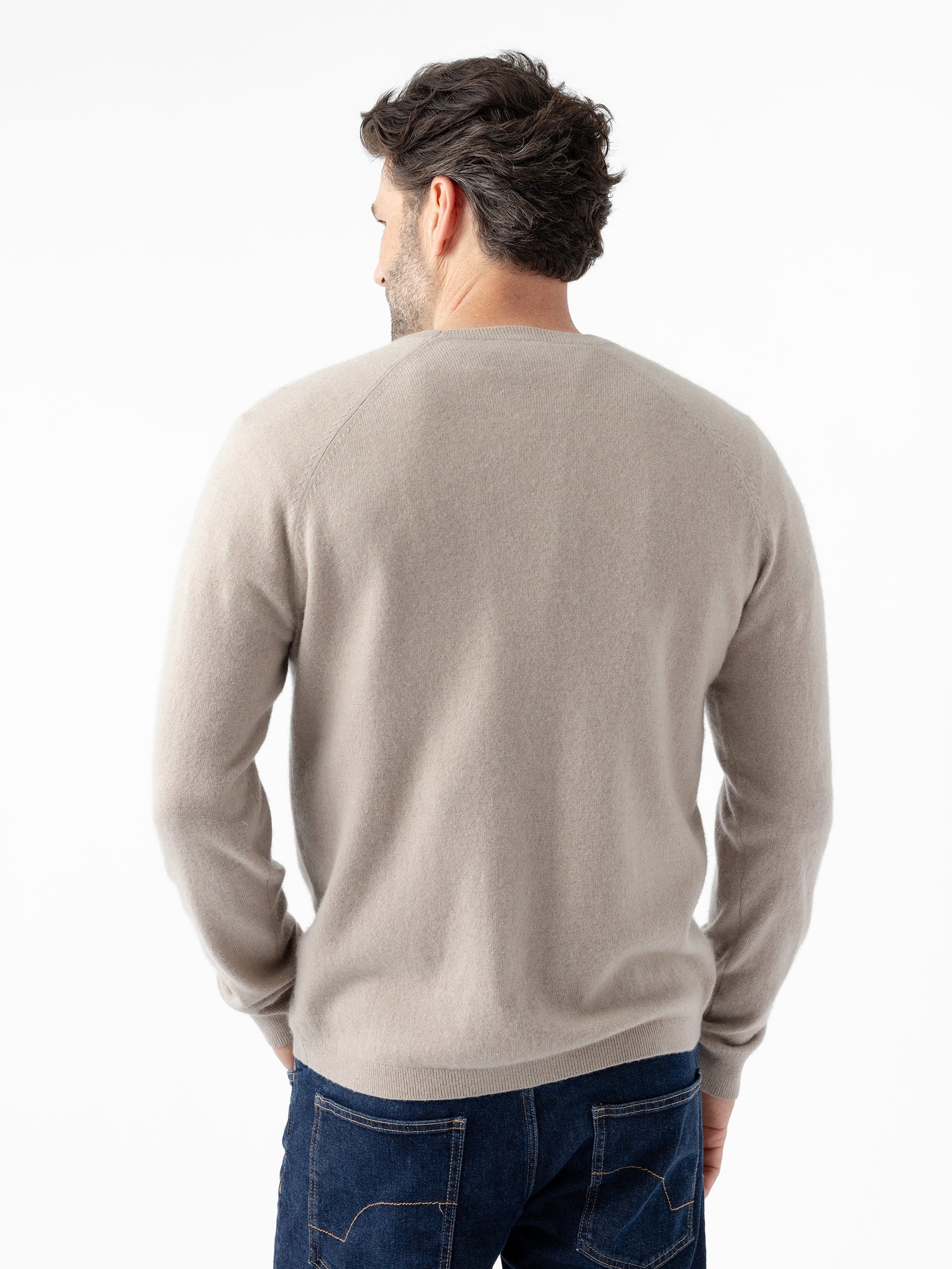 A man with short dark hair is facing away from the camera. He is wearing a Men's Crewneck Sweater in light beige by Cozy Earth and dark blue jeans. The background is white and uncluttered. 