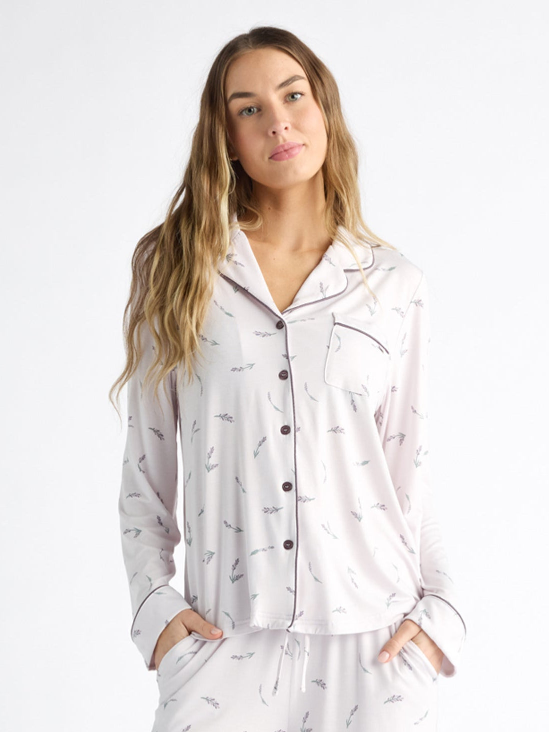 A person wearing Cozy Earth Women's Bamboo Stretch-Knit Long Sleeve Pajama Top, featuring a collar, buttons, and chest pocket, stands relaxed against a white background with long hair and hands in pockets. The light-colored top has a subtle print. 