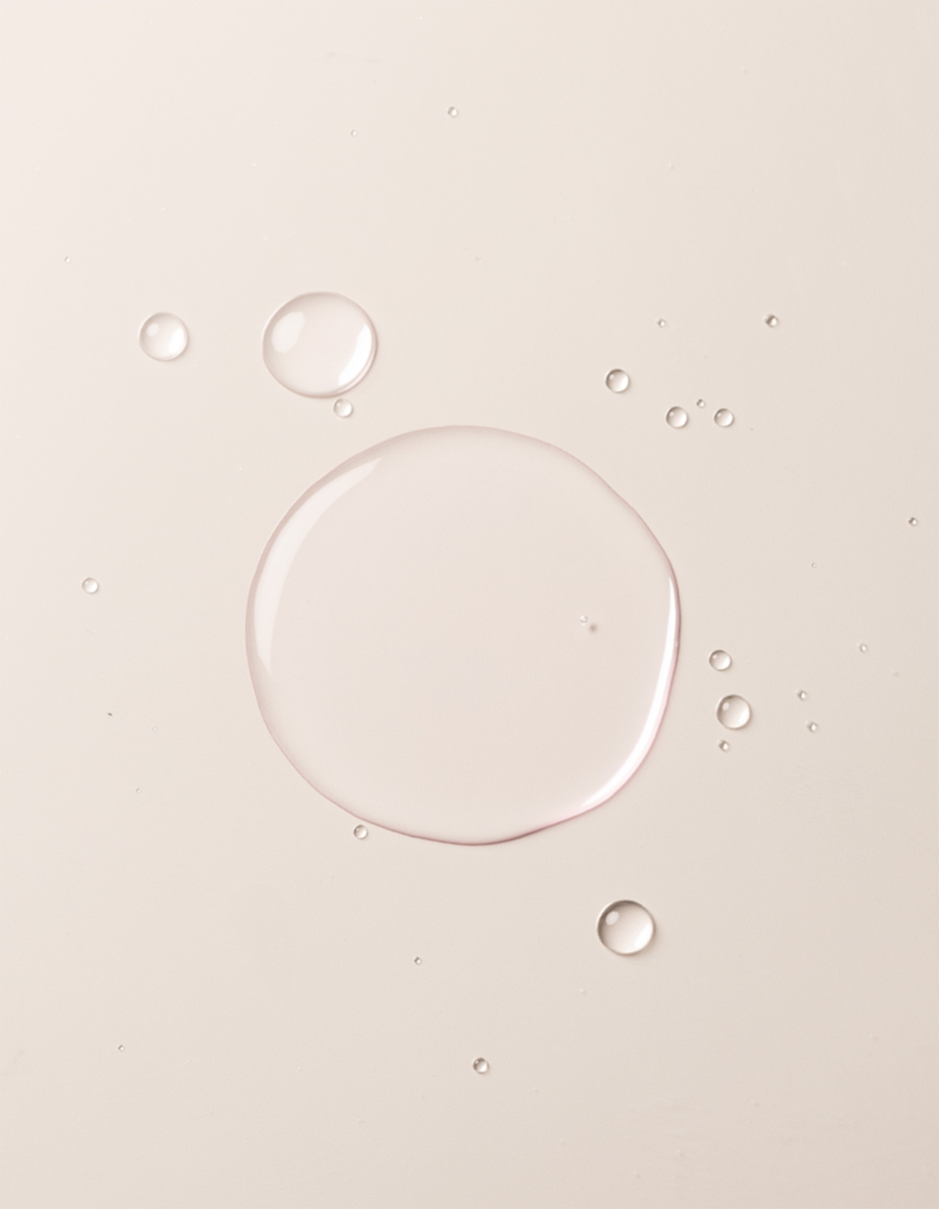 A close-up image showcases translucent liquid droplets of Cozy Earth's Balancing Face Mist on a smooth, off-white surface. The droplets vary in size, with one large central droplet surrounded by several smaller ones, creating a clean and minimalistic appearance.