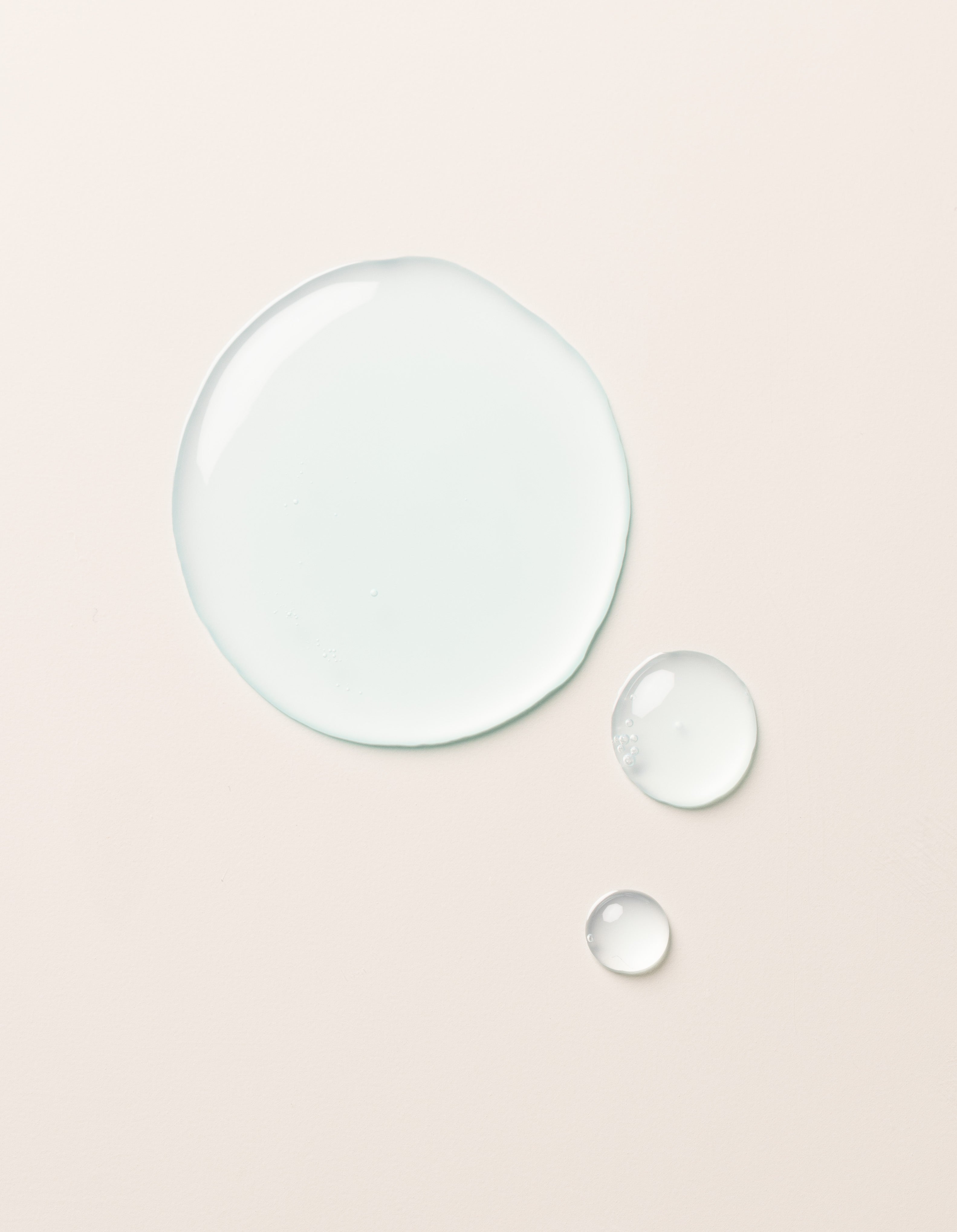 A large clear droplet and two smaller droplets of Cozy Earth Hydrating Serum rest on a smooth, light beige surface. The glossy, semi-transparent droplets reflect light slightly on their surfaces.