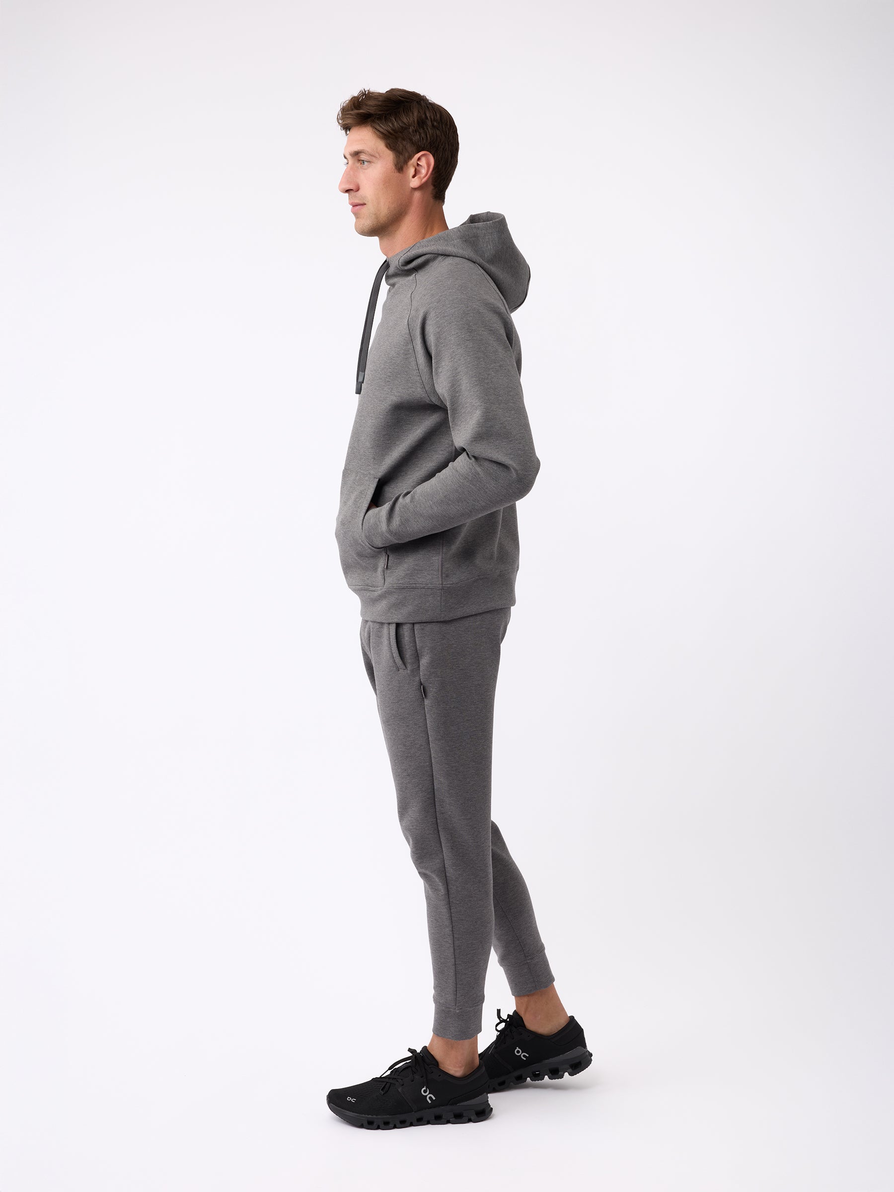 A person stands in profile against a white background, wearing the Cozy Earth Men's StretchTech Hoodie and matching joggers with black sneakers. They have their hands in the hoodie pockets and are looking forward. |Color:Coal Heather