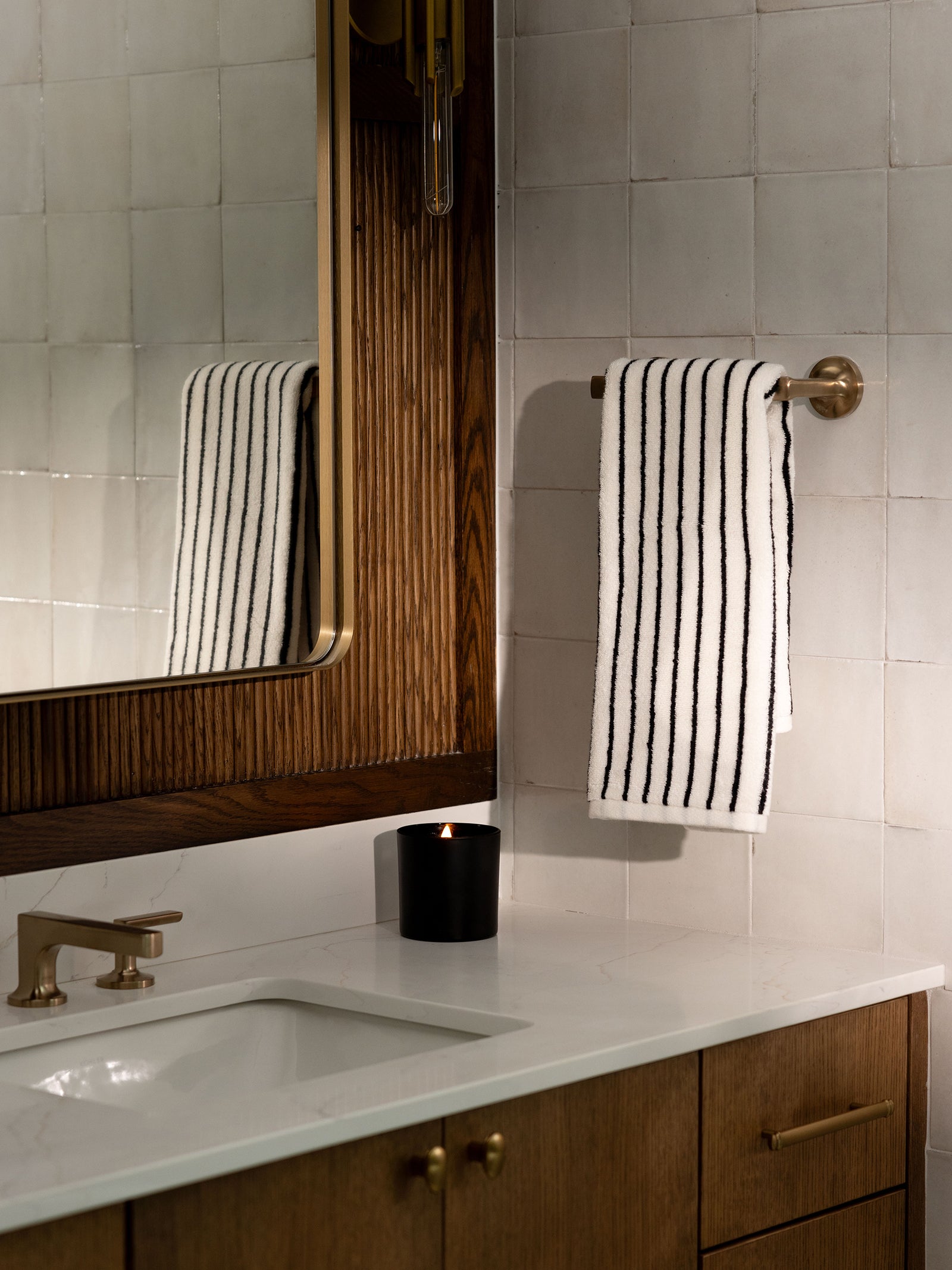 A bathroom scene featuring a white countertop with a sink, brass faucet, and wooden cabinets. A black candle and Luxe Hand Towels by Cozy Earth, hanging on a brass towel bar, are also present. The background consists of white tiled walls and a large wooden-framed mirror. 