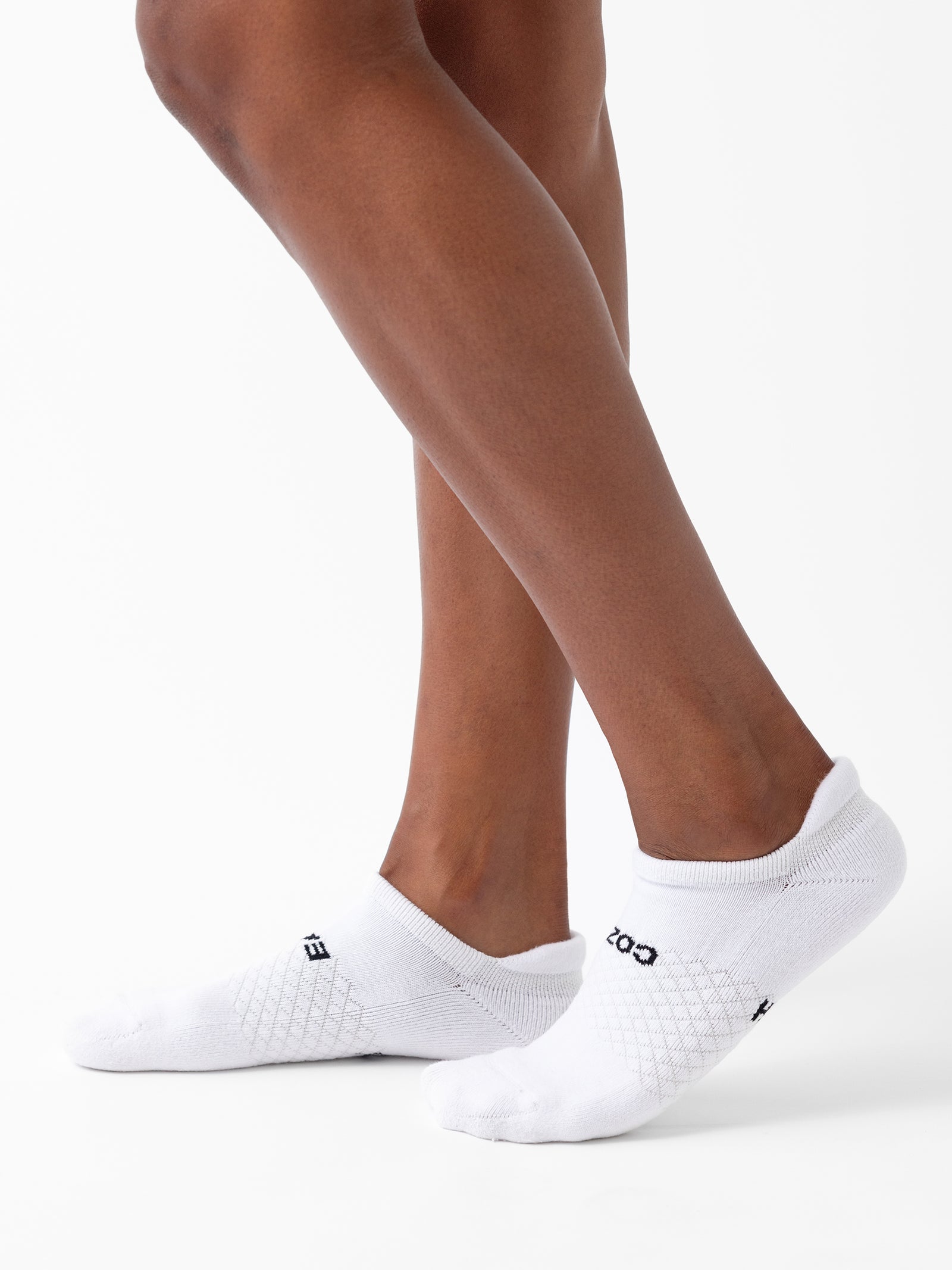 A person models Cozy Earth's Essential Ankle Sock 8-Pack against a plain white background, with one foot slightly raised and legs visible from the knees down. The white socks feature a textured pattern and text detail. 