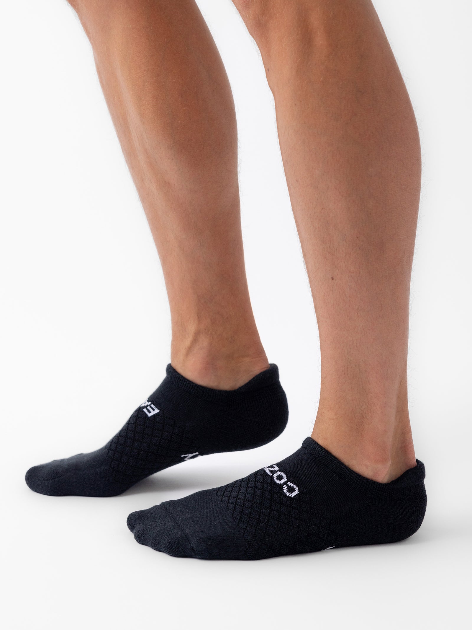 A person stands on tiptoe wearing Cozy Earth Essential Ankle Socks from the 6-Pack, featuring a textured pattern and white lettering, shown against a white background. 
