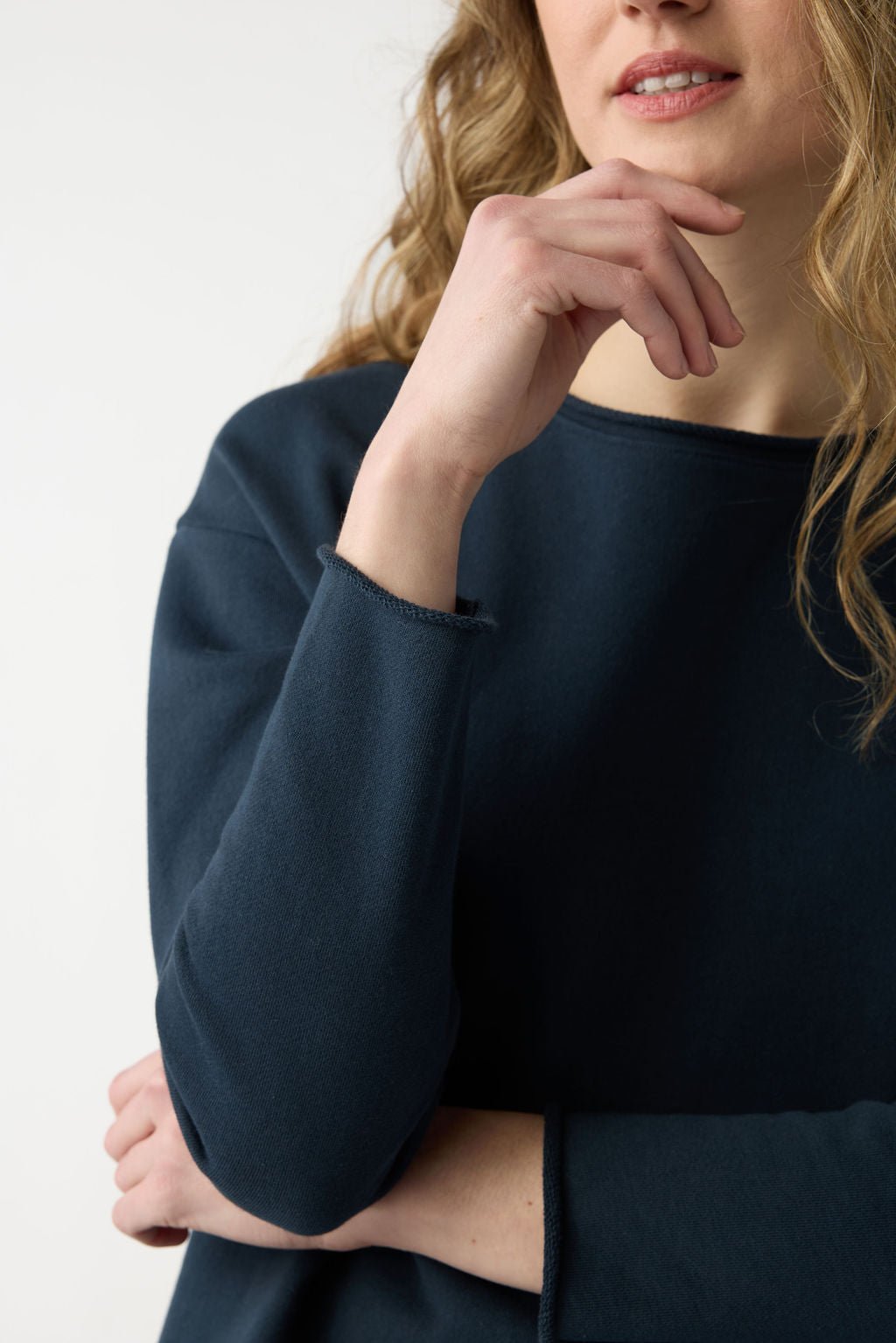 A person with wavy blonde hair wears the Cozy Earth Women's Chelsea Pullover in dark blue, posed with their left hand on their chin and right arm crossed against a neutral background.