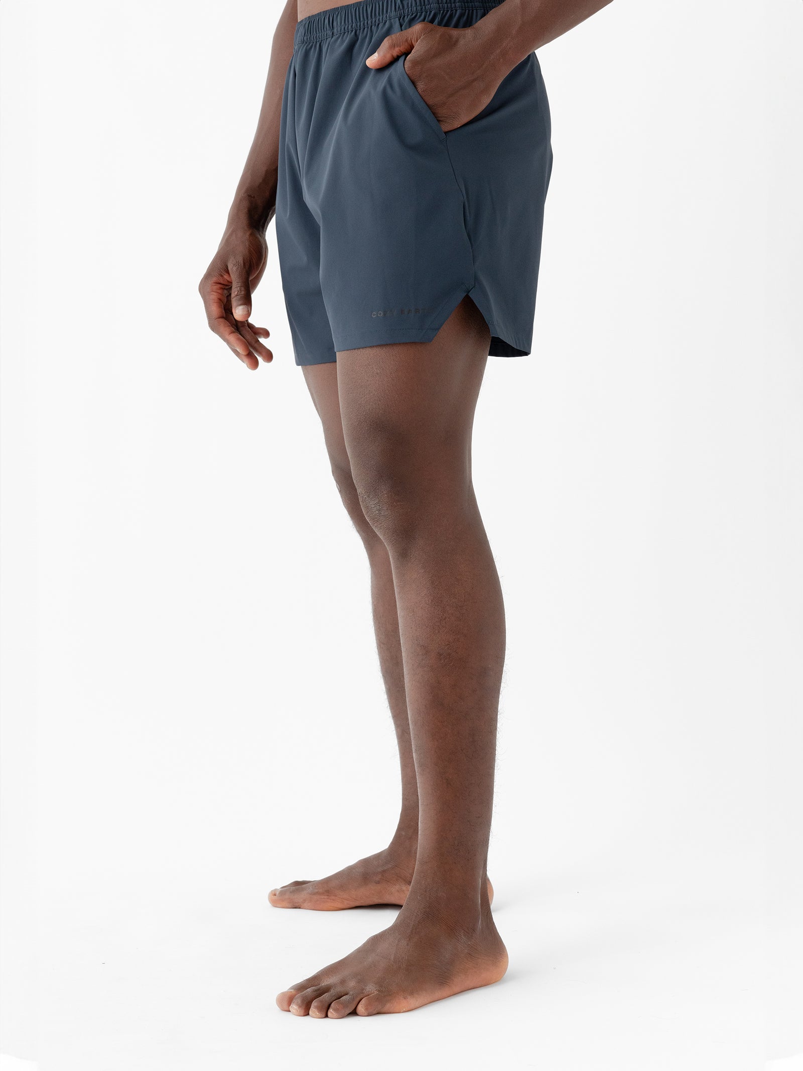 A person stands barefoot against a white background, wearing dark blue Men's Performance Sleep Shorts by Cozy Earth. Their hands are relaxed by their sides, with the right hand placed inside a pocket. The shorts feature a subtle logo near the bottom hem on the left side. 