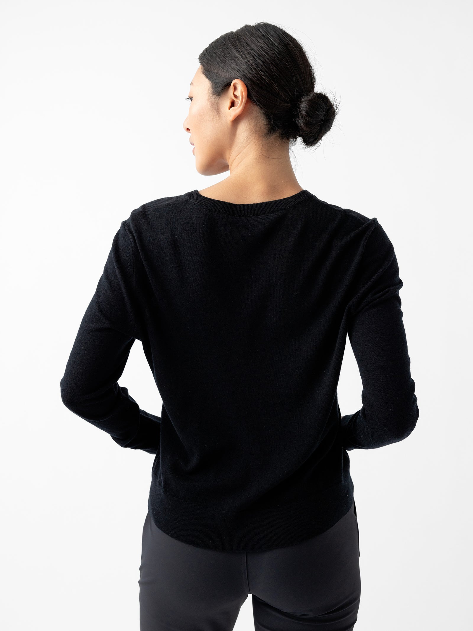 A person with dark hair tied in a low bun is wearing a black Women's AirKnit V-Neck Sweater from Cozy Earth and dark pants. They are standing facing away from the camera against a plain white background, with their hands placed on their hips. 