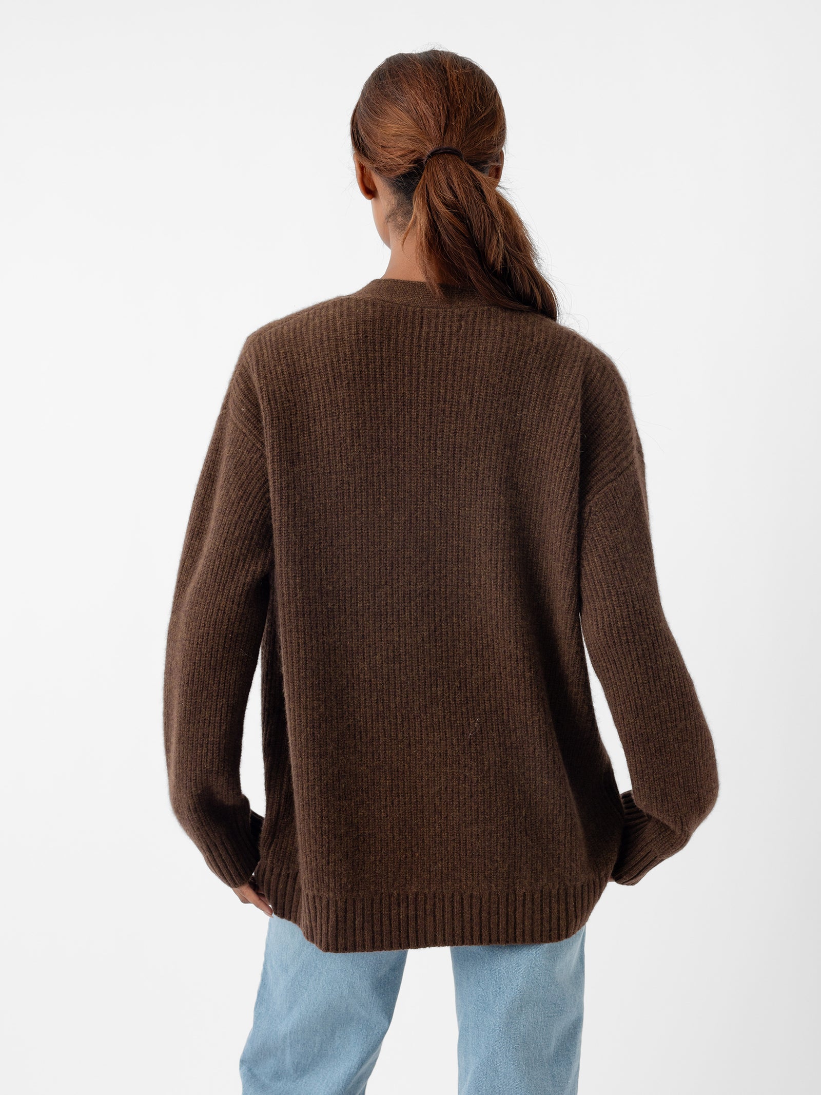 Rear view of a person with brown hair tied in a ponytail, wearing the Cozy Earth Oversized Sunday Cashmere Cardigan and light blue jeans, standing against a plain white background. 