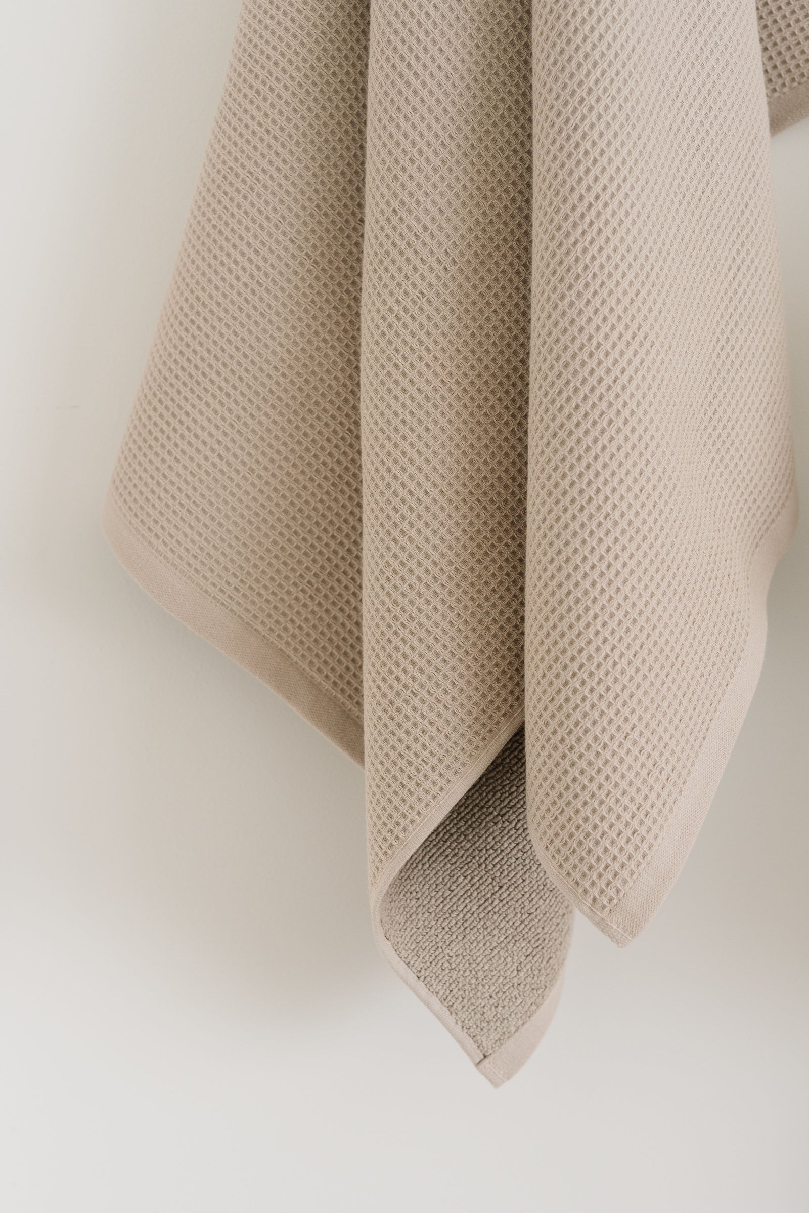 Waffle Bath Towel in the color Sand. Photo of Sand Waffle Bath Towel taken hanging from a towel hook in a white bathroom. The upper half of the towel and towel hook is not seen. 