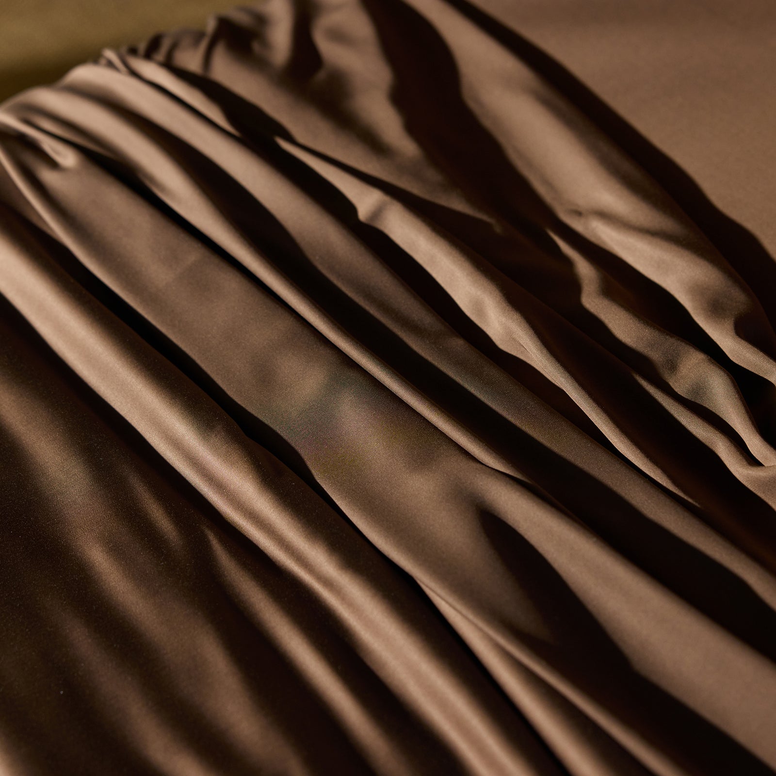 Close-up image of gathered and draped Bamboo Sheet Set by Cozy Earth in a rich brown color, creating soft, flowing folds with highlights and shadow play. The texture appears smooth and slightly shiny, suggesting a luxurious and elegant material. 