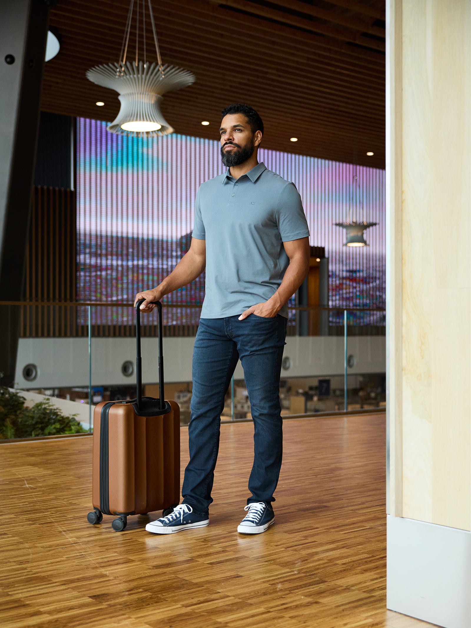 A man stands indoors with a hand on the extended handle of a bronze-colored rolling suitcase. He is wearing Cozy Earth's Men's Everyday Polo in light gray, paired with dark jeans and black sneakers. The background features modern wooden architecture and a large digital screen. 