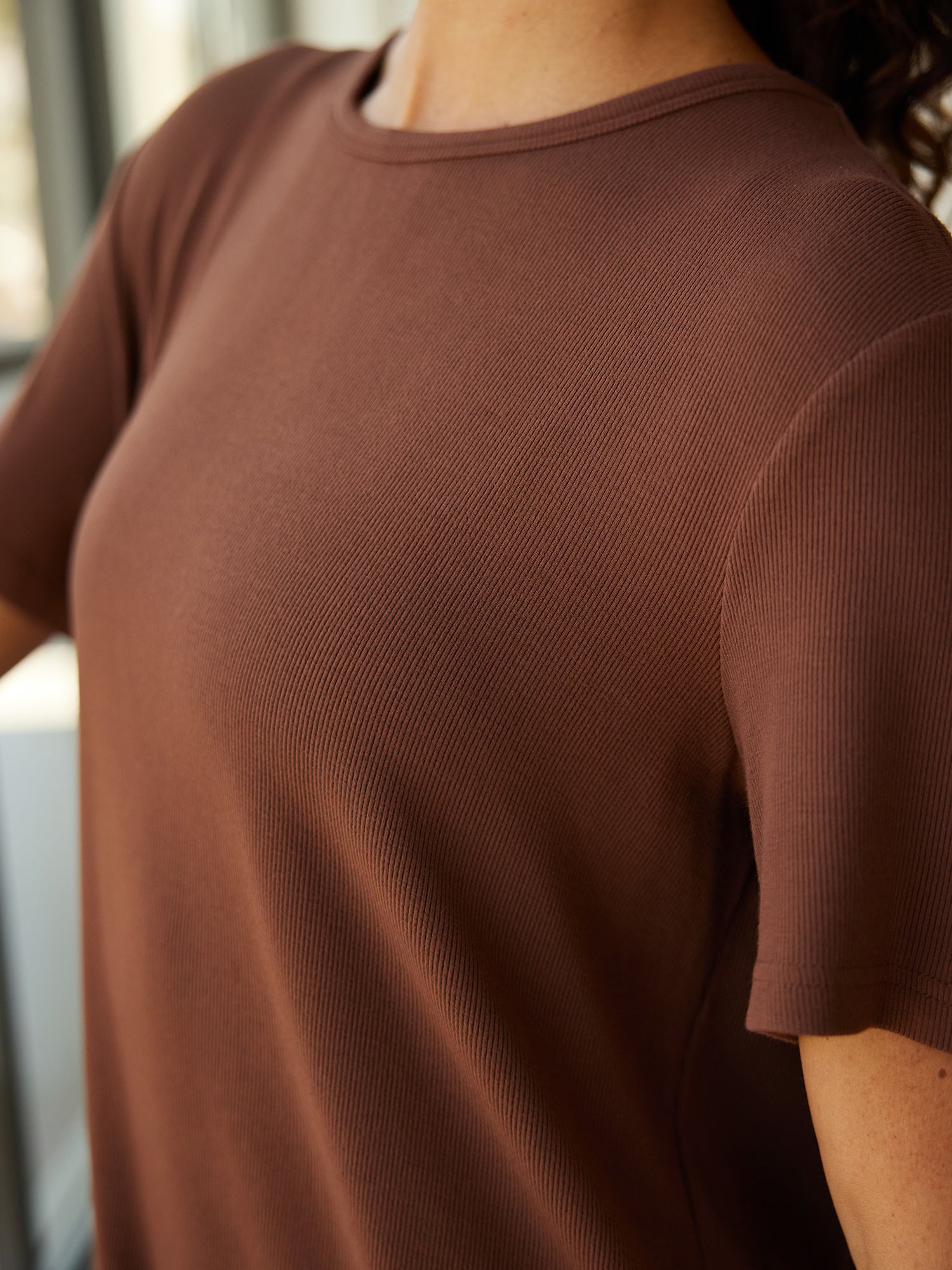 Close-up image of a person wearing Cozy Earth's Women’s Bamboo Rib-Knit Boyfriend Sleep Dress in brown. The focus is on the upper part of the sleep dress and the right shoulder, with the person's head not visible. The background appears to be indoors with blurred windows. 