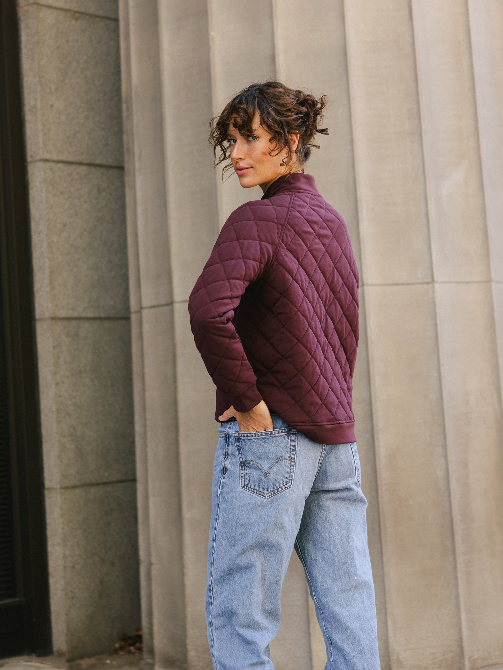 A person with curly hair is standing with their back turned, looking over their shoulder. They are wearing the Cozy Earth's Women's Ultra-Soft Bamboo Quilted Snap Pullover in maroon and light blue jeans. The background features a gray, vertically ribbed concrete wall. 