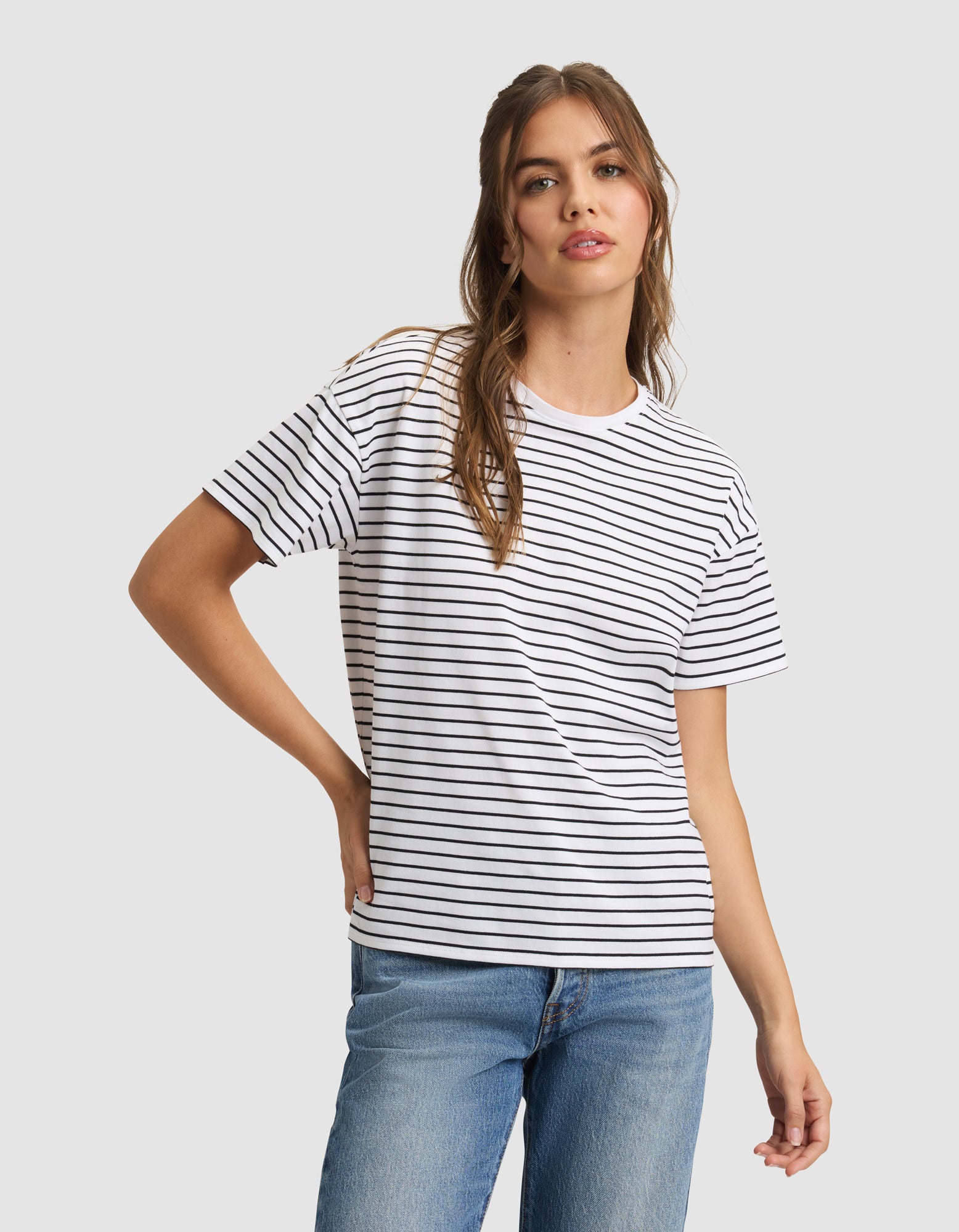 A woman models the Cozy Earth Women's All Day Tee—a white t-shirt with black stripes—paired with blue jeans, standing against a light gray background and looking at the camera, one hand on her hip. 
