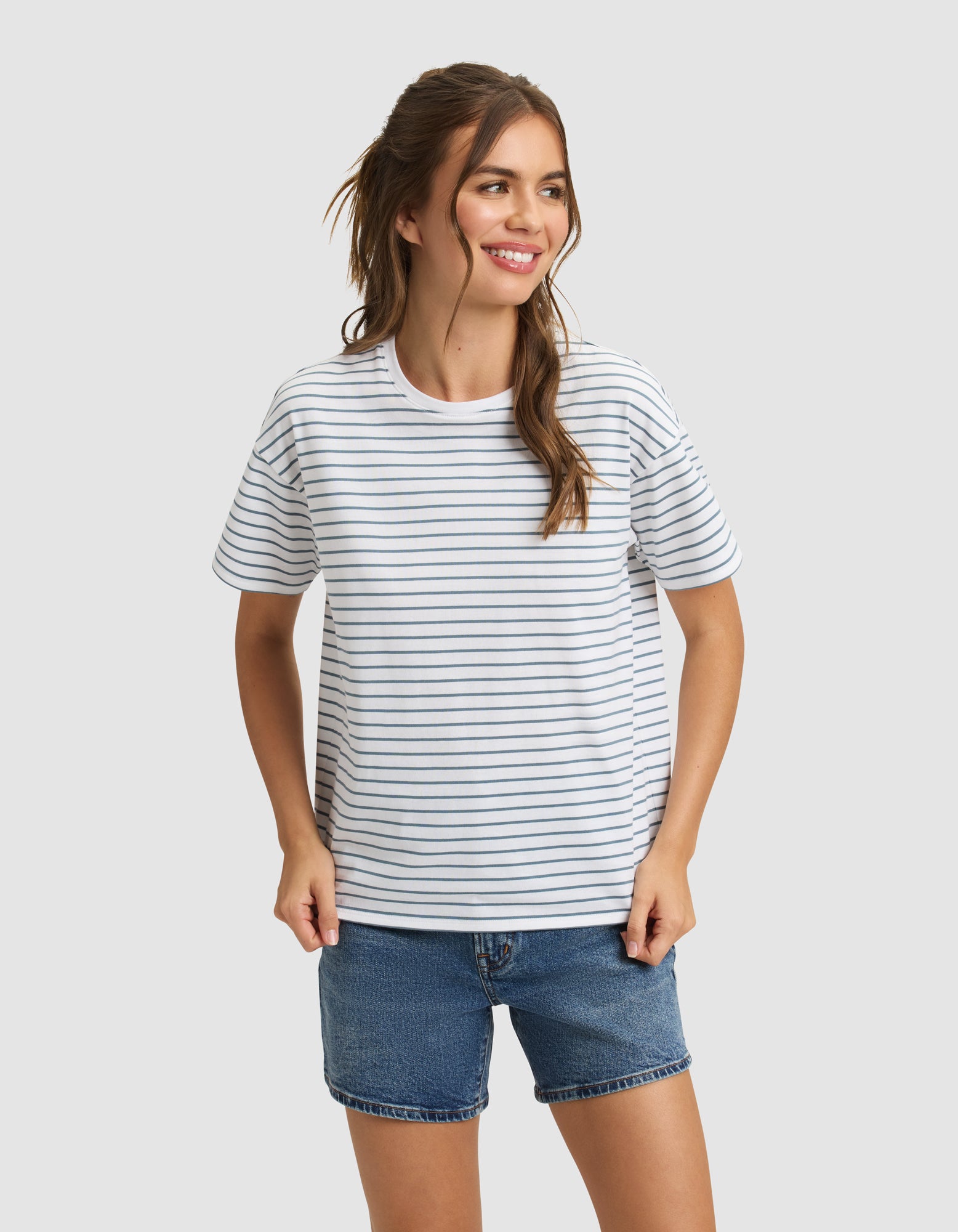A woman with long brown hair in a ponytail wears the Cozy Earth Women's All Day Tee, featuring thin black stripes, and blue denim shorts. She smiles and looks to the side against a plain light gray background. 