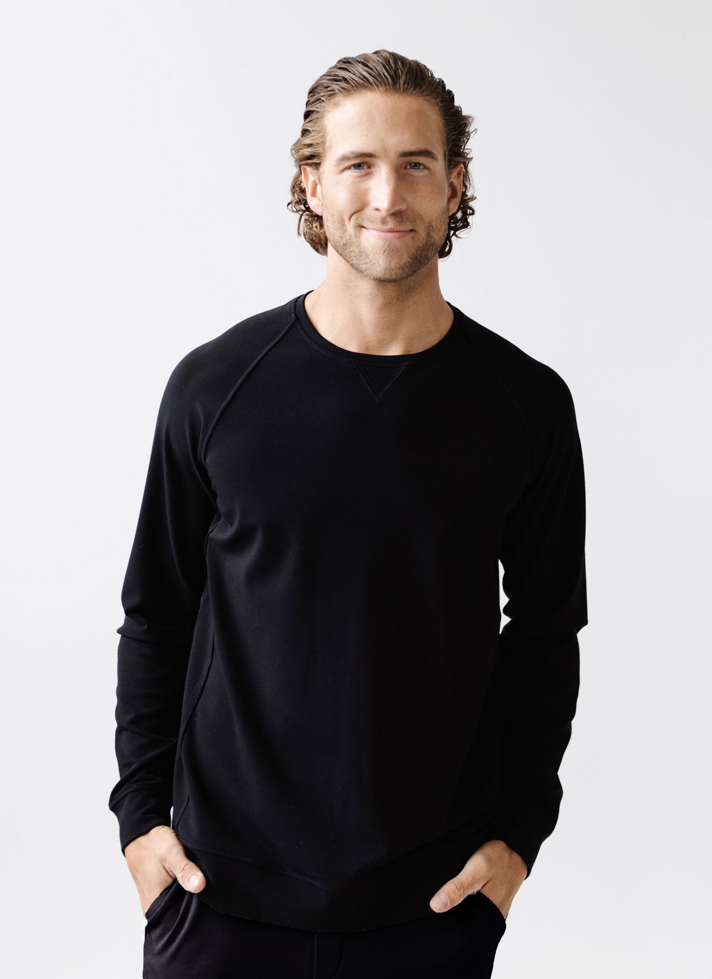 A person with wavy hair smiles with hands in their pockets, wearing a black Men's Bamboo Jogger Set by Cozy Earth against a plain white background. |Color:Black