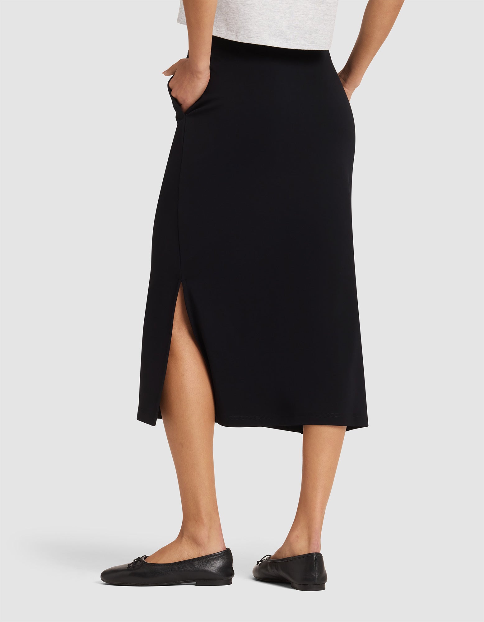 Model wears Cozy Earth's Women's Brushed Bamboo Midi Skirt, featuring a side slit, paired with black flats and a light top. She stands hands in pockets, showcasing the skirt’s back and side views. |Color:Black