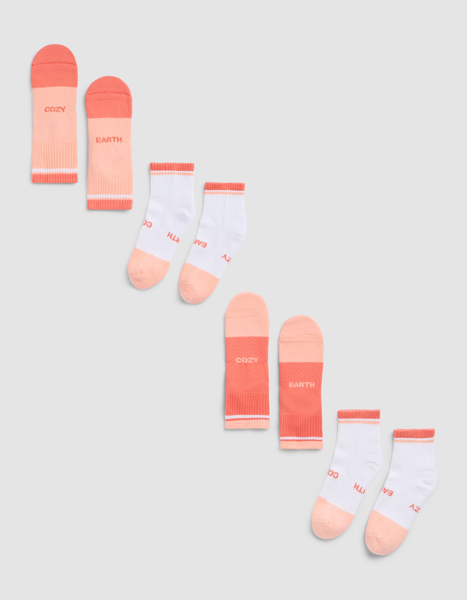 The Cozy Earth Essential Quarter Sock 4-Pack is shown on a gray background, featuring peach and pink pairs with "COZY" and "EARTH" text, plus white pairs accented in peach and pink with similar lettering. 