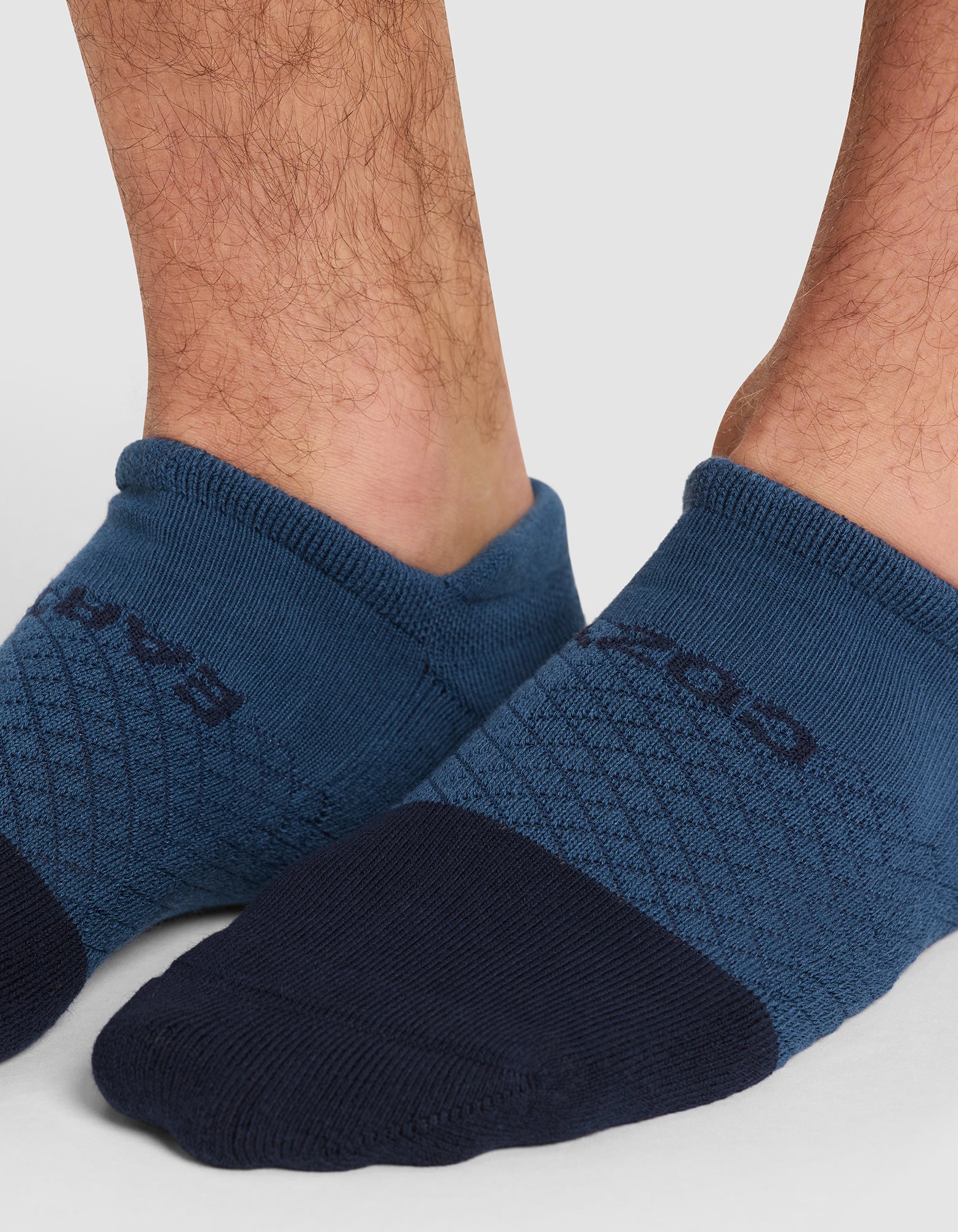 Close-up of a person's lower legs wearing Cozy Earth Essential Ankle Socks in blue and navy, featuring a textured pattern and partially visible logo. The background is plain and light-colored. 