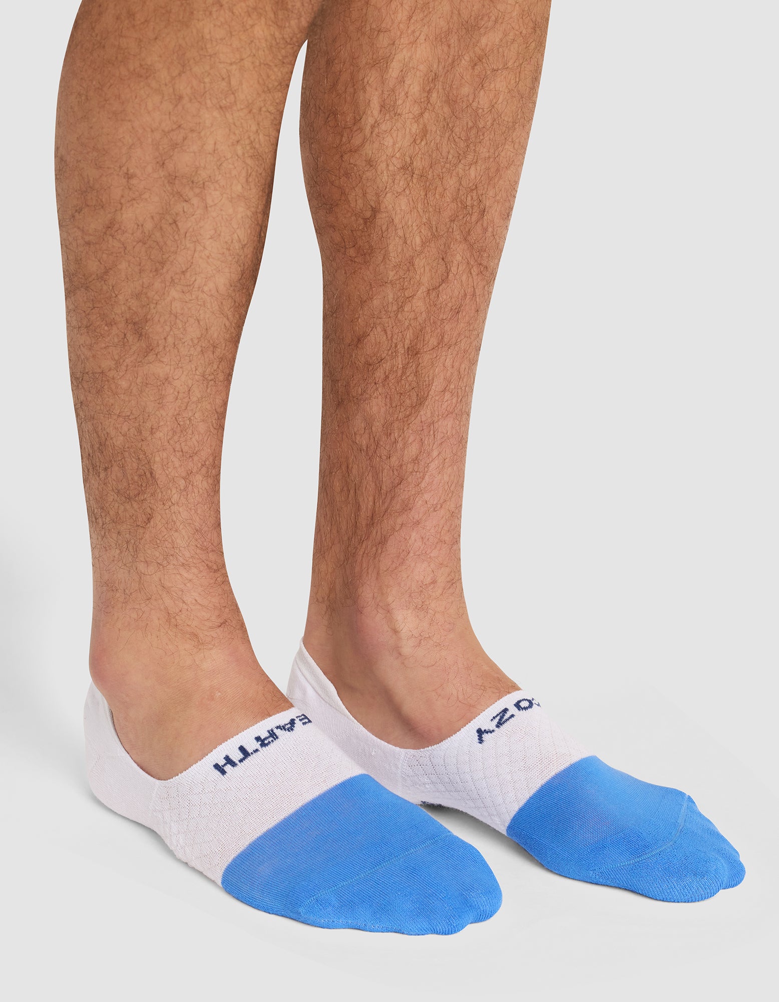 A person wearing Cozy Earth's Essential No-Show Sock in white and blue with visible text near the ankle, standing on a white background. 