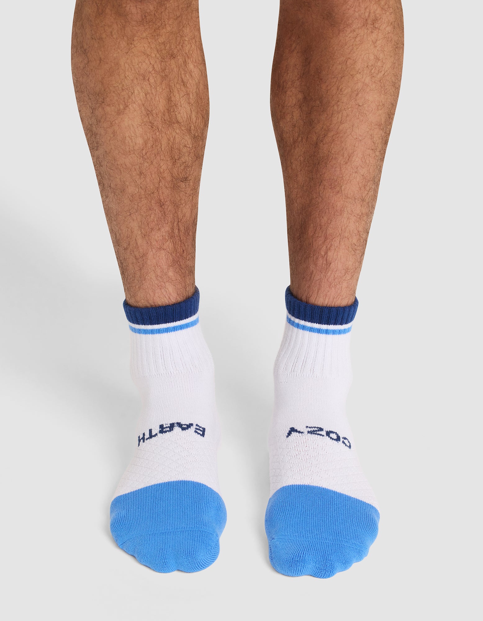 A person stands on a light surface wearing Cozy Earth Essential Quarter Socks in white with blue toes and cuffs and "EARTH" and "COZY" printed in dark blue on top. 
