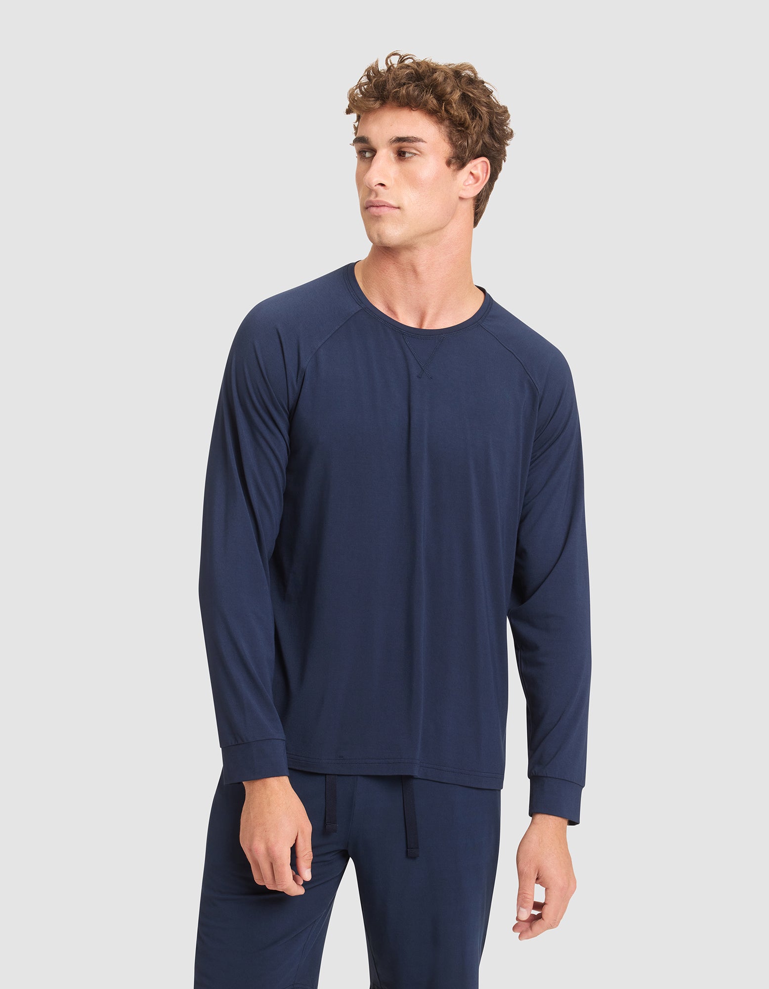 A young man with curly hair wears Cozy Earth's Men's Bamboo Stretch-Knit Long Sleeve in navy blue, paired with matching pants, standing against a plain light gray background and looking slightly to his left.  |Color:Navy