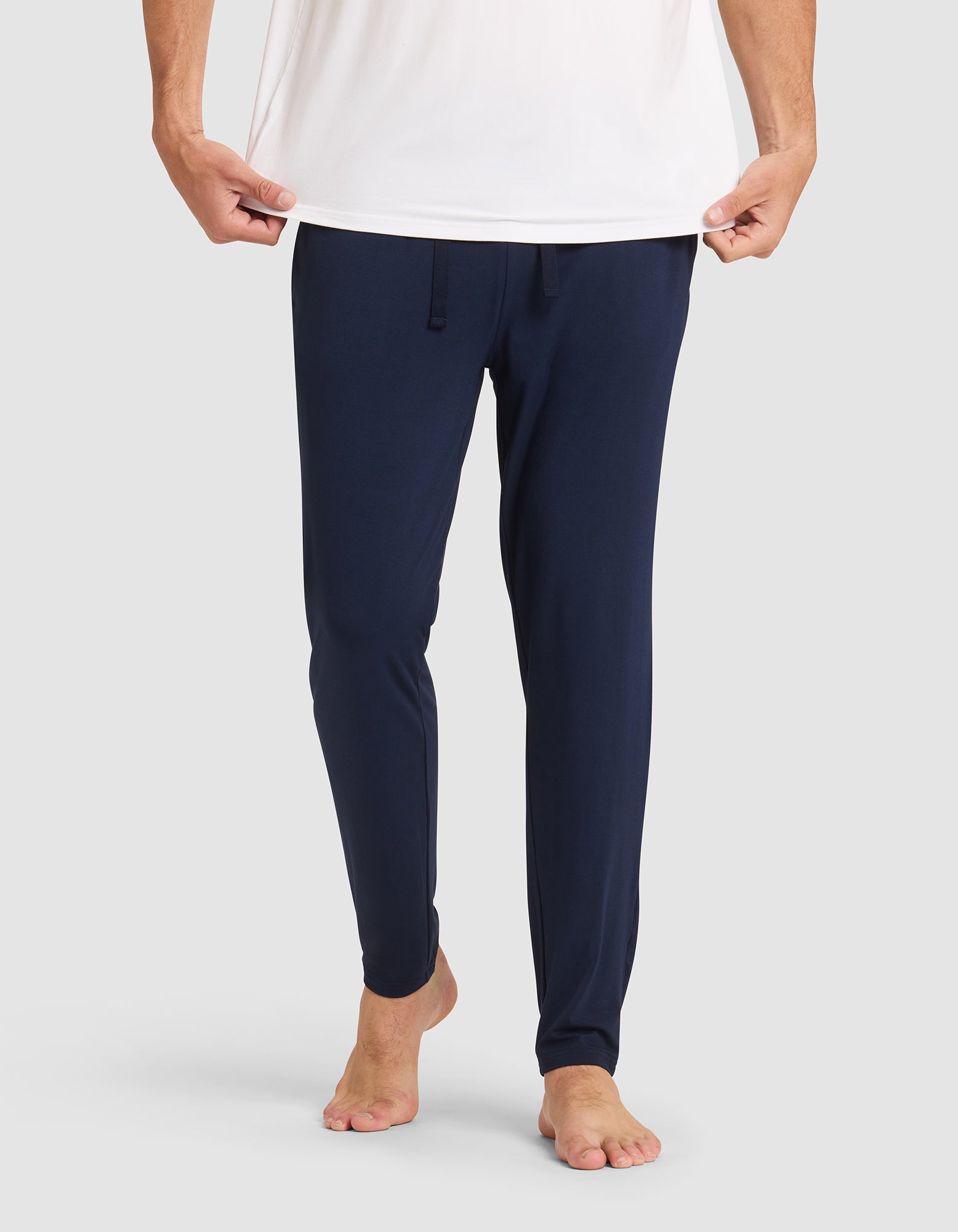 Only the lower half of a person is shown against a plain light background, barefoot and wearing Cozy Earth's Men's Bamboo Stretch-Knit Pajama Pant in navy blue with a drawstring waist and a white shirt, holding the shirt's hem. |Color:Navy