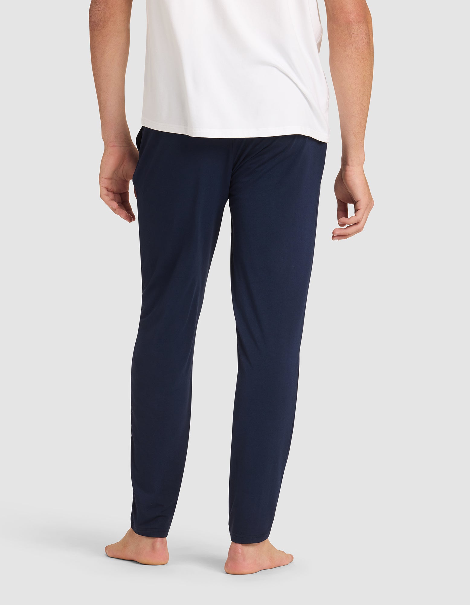 A person stands barefoot, shown from the back, wearing a white shirt and Cozy Earth Men's Bamboo Stretch-Knit Pajama Pant in dark blue against a plain light background. |Color:Navy