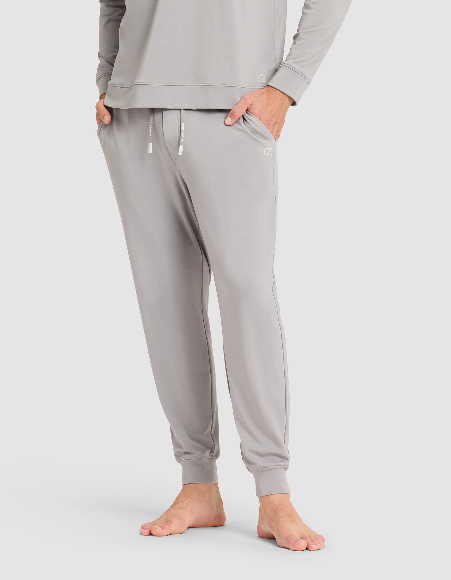 A person stands barefoot against a plain light background, wearing Cozy Earth's Men's Brushed Bamboo Jogger Pant in light gray with hands in pockets, paired with a matching long-sleeve top. |Color:Stone