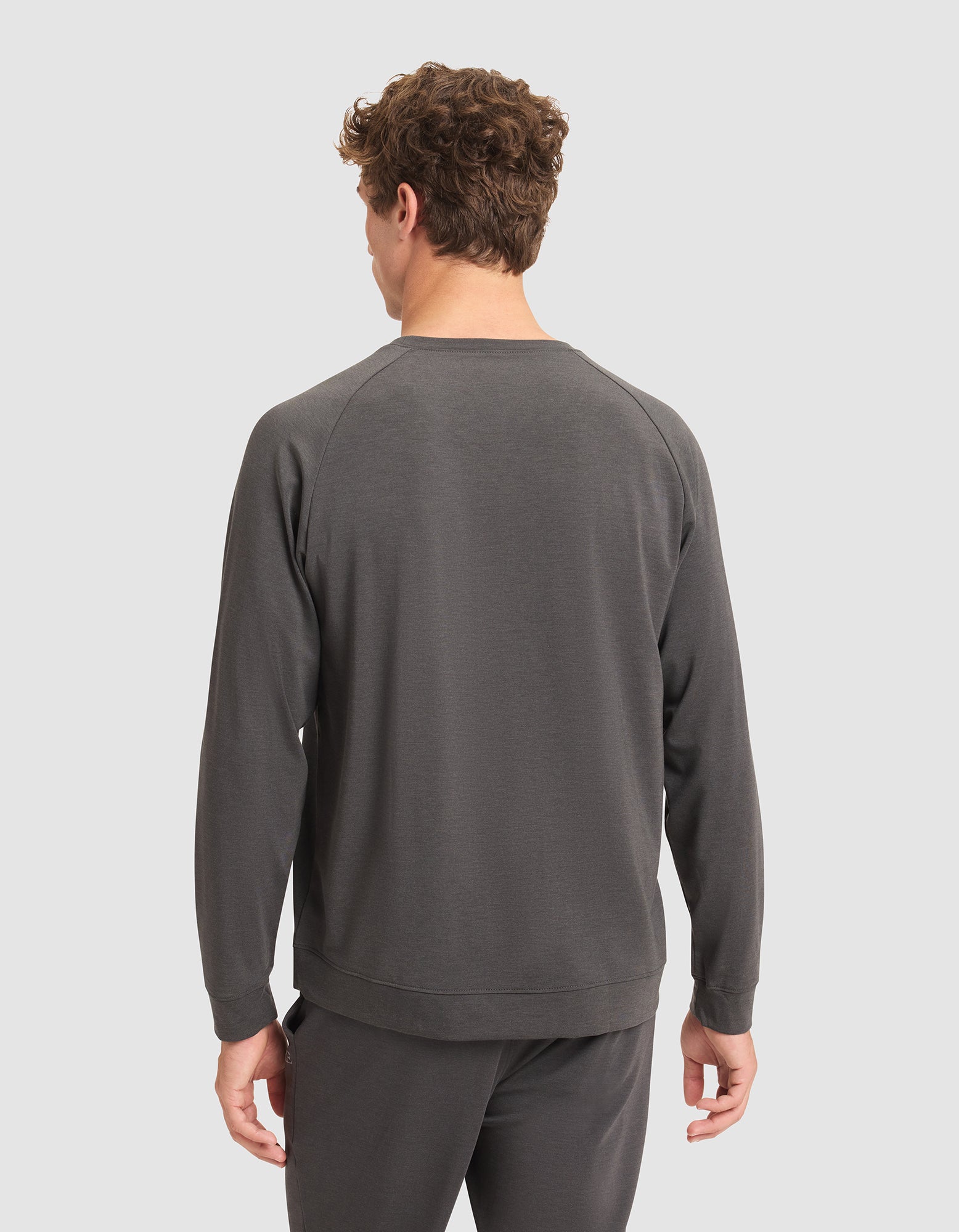 A man with short curly hair faces away from the camera, wearing a dark gray Cozy Earth Men's Brushed Bamboo Pullover Crew and matching pants against a plain light gray background. 