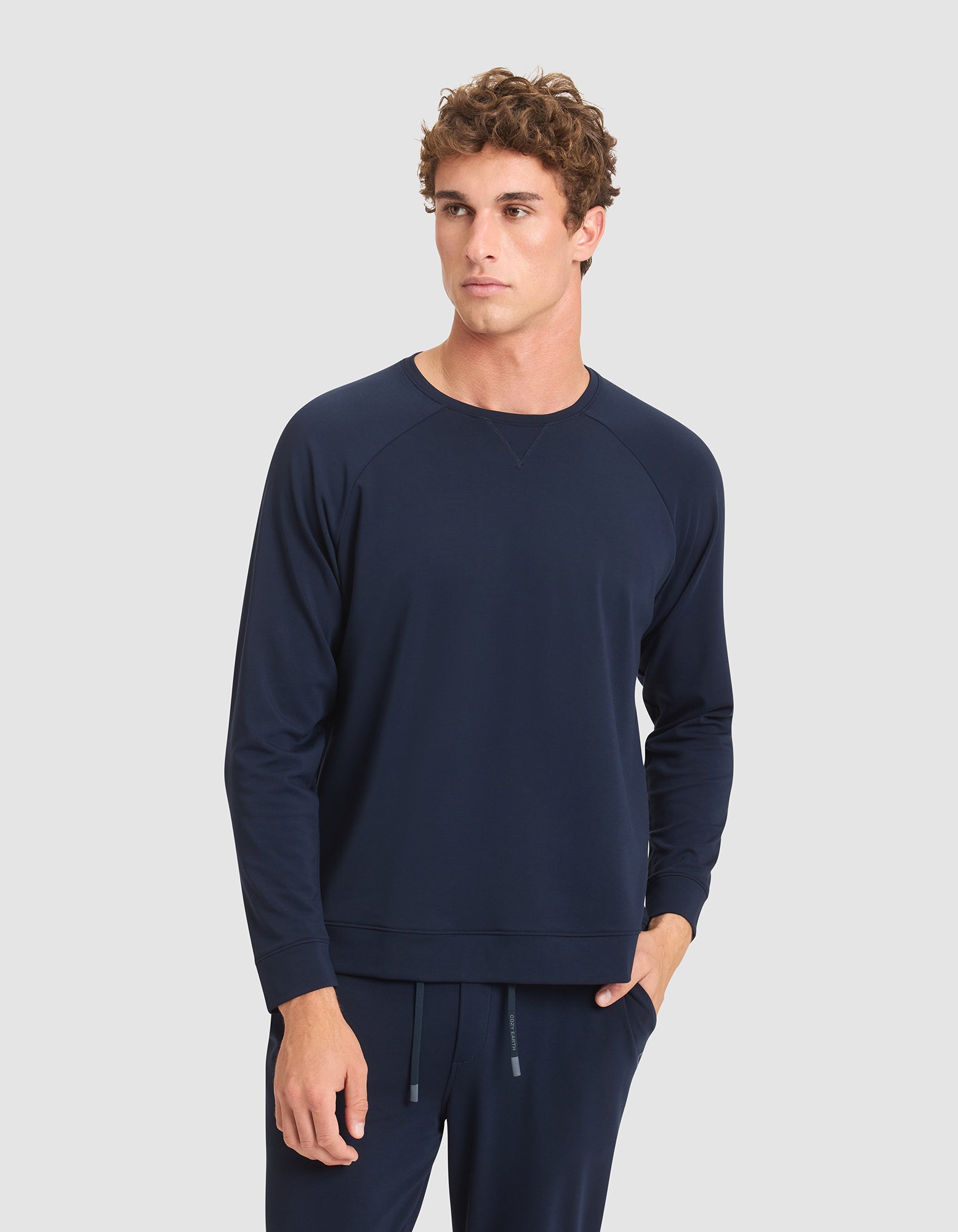 A man wearing the Cozy Earth Men's Brushed Bamboo Pullover Crew in navy blue stands against a light gray background, one hand in his pocket and a neutral expression on his face. 